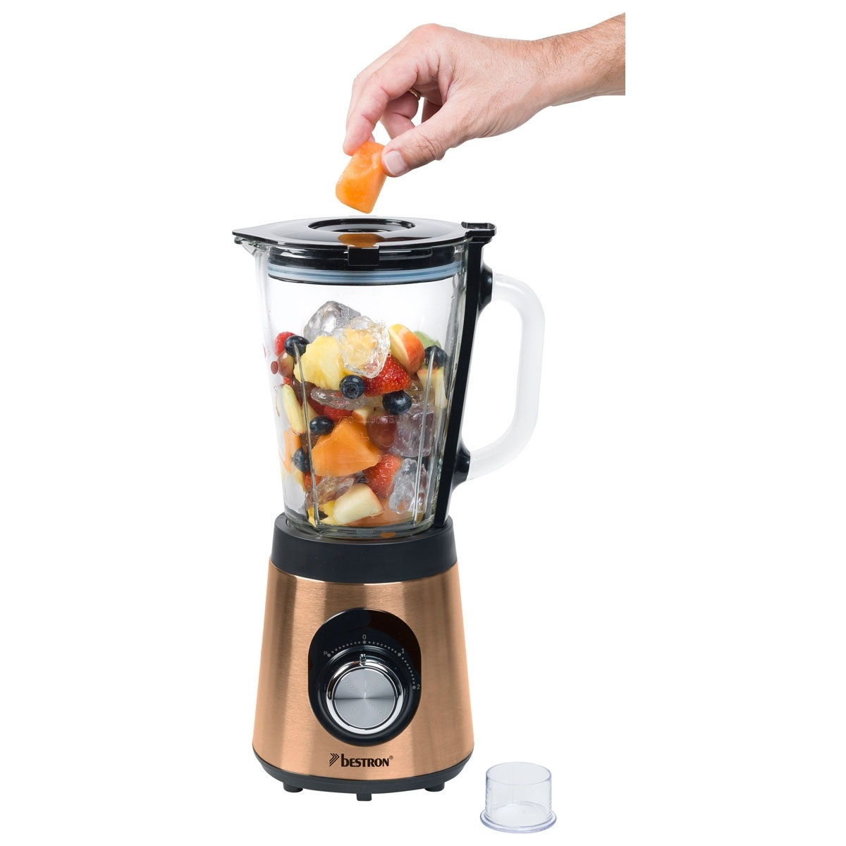 Eine Hand fügt einem Mixer, der mit Eis und Obst gefüllt ist, ein Stück orangefarbenes Obst hinzu. Der Mixer ist kupferfarben.