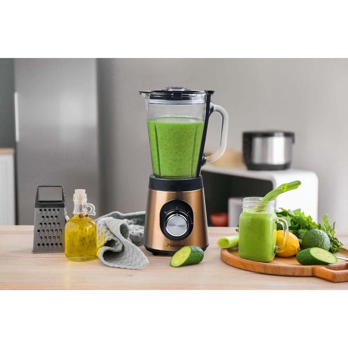 Ein Mixer mit grünem Smoothie, Zutaten und einem Glas Smoothie stehen auf einem Holztisch. Der Hintergrund hat einen Kühlschrank und ein weißes Regal.