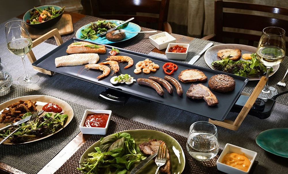 Ein Tisch ist mit Essen gedeckt: gegrilltes Fleisch, Meeresfrüchte, Salate und Saucen. Weingläser sind vorhanden. Ein Elektrogrill steht im Zentrum.