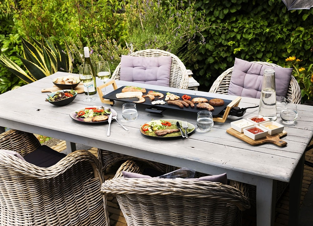 Szene im Freien mit gegrilltem Essen, Wein und Salaten auf einem Holztisch. Korbstühle umgeben den Tisch, Garten im Hintergrund.
