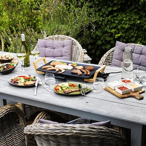 Szene im Freien. Ein Tisch mit Essen, Getränken und einem Grill. Das Essen umfasst Fleisch, Salat und Saucen. Korbstühle umgeben.