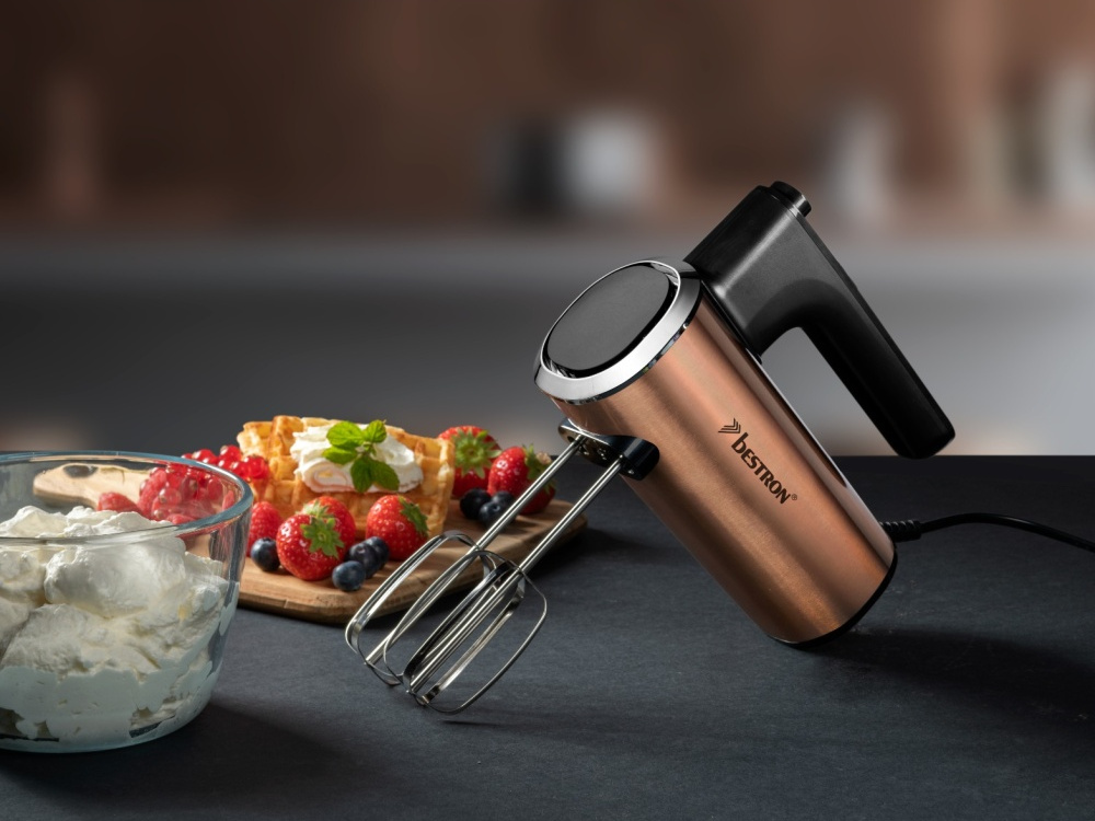 Ein kupferfarbener Handmixer neben Waffeln, Beeren und Schlagsahne. Der Mixer steht auf dunklem Untergrund, mit verschwommenem Hintergrund.