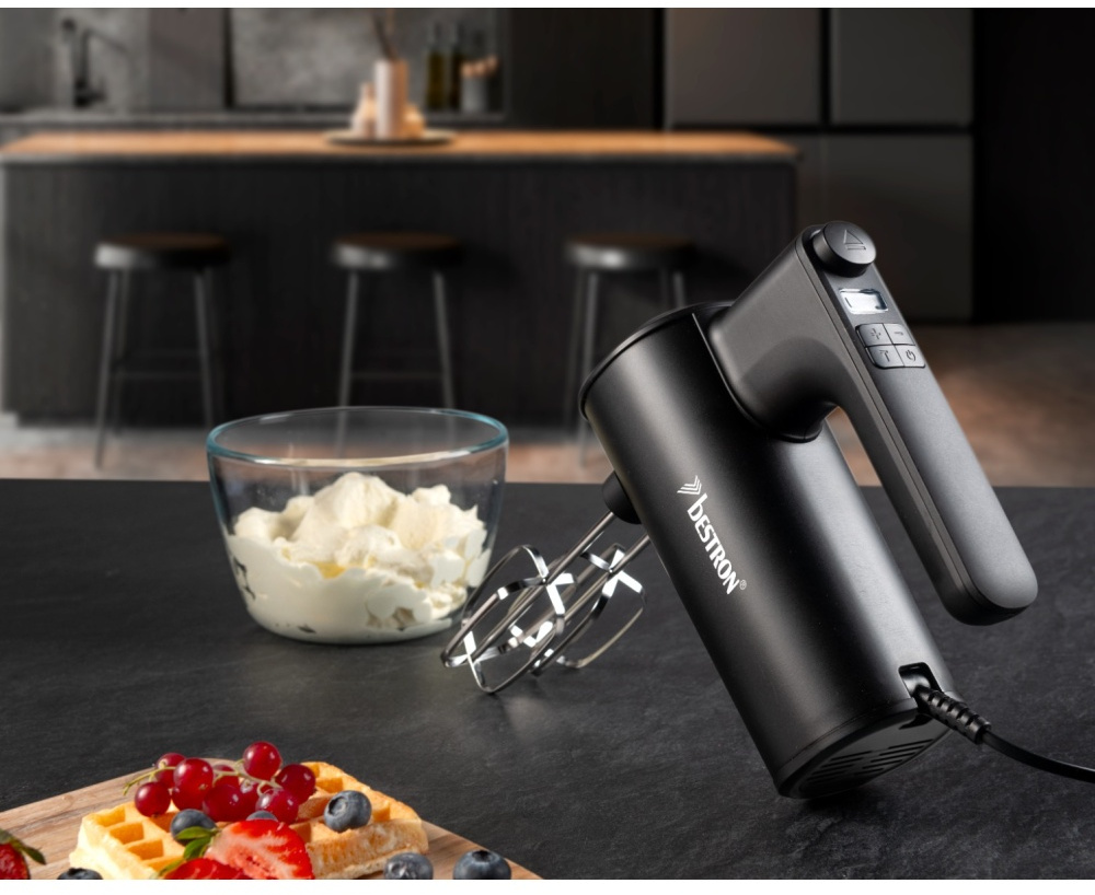 Ein schwarzer Handmixer steht auf einem Tisch. Eine Schüssel mit Sahne, ein Teller mit Waffeln und Früchten stehen ebenfalls auf dem Tisch, Küche im Hintergrund.