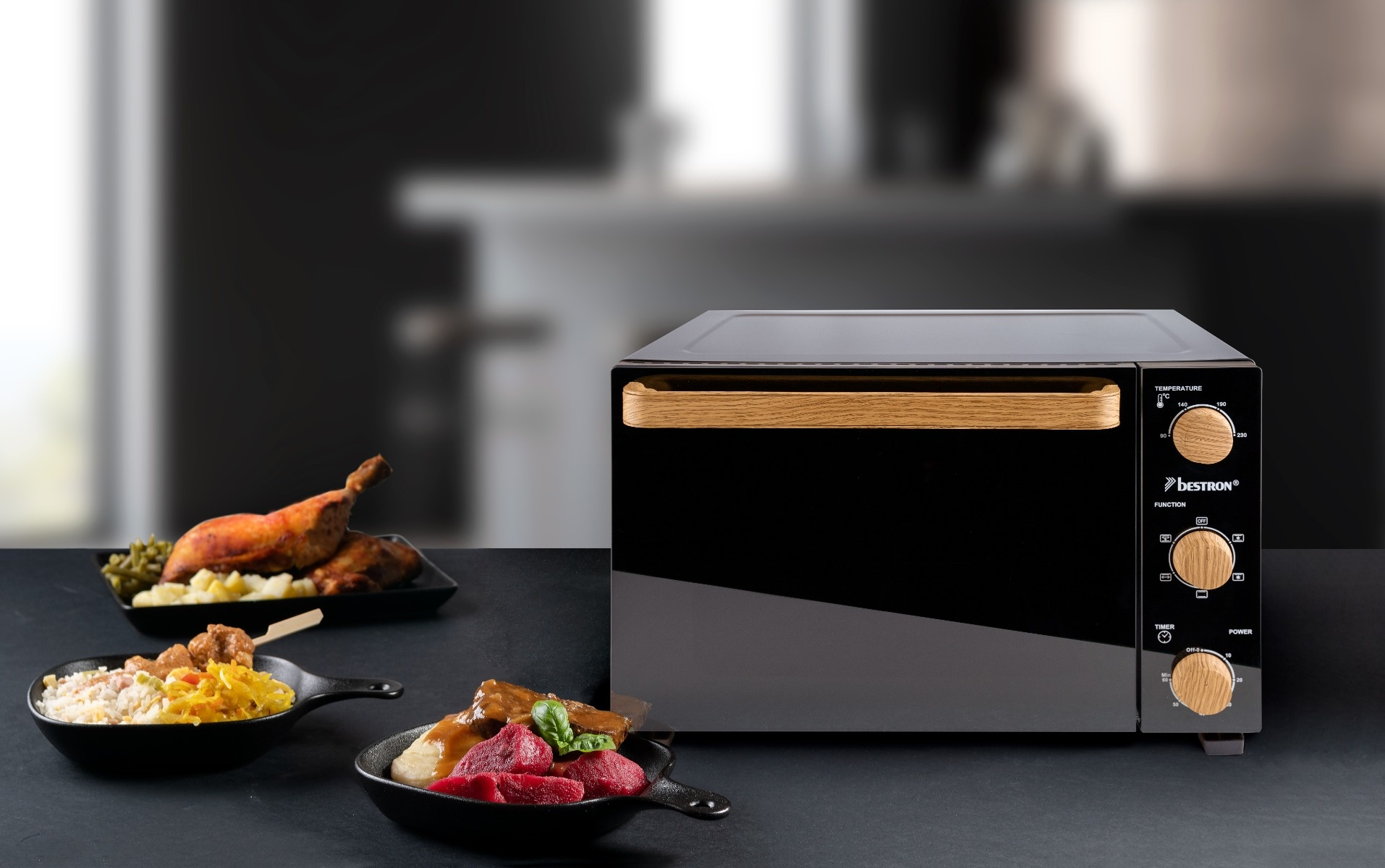 Schwarzer Ofen mit Holzgriff und Bedienknöpfen, begleitet von gekochtem Essen in schwarzen Pfannen. Der Hintergrund ist unscharf.