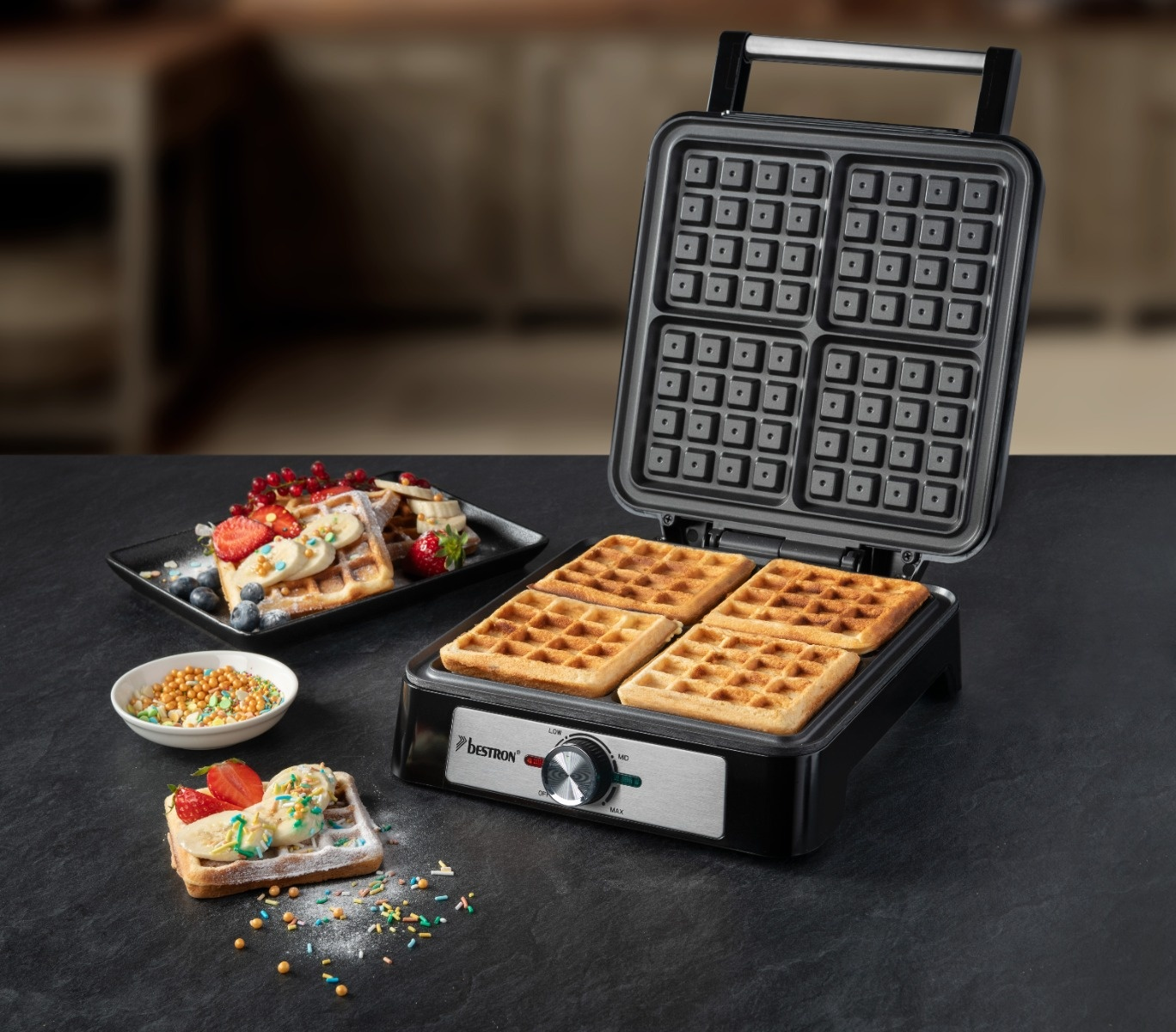 Waffeleisen mit fertigen Waffeln, servierten Waffeln mit Belag und einer Schüssel mit Streuseln auf schwarzer Oberfläche.