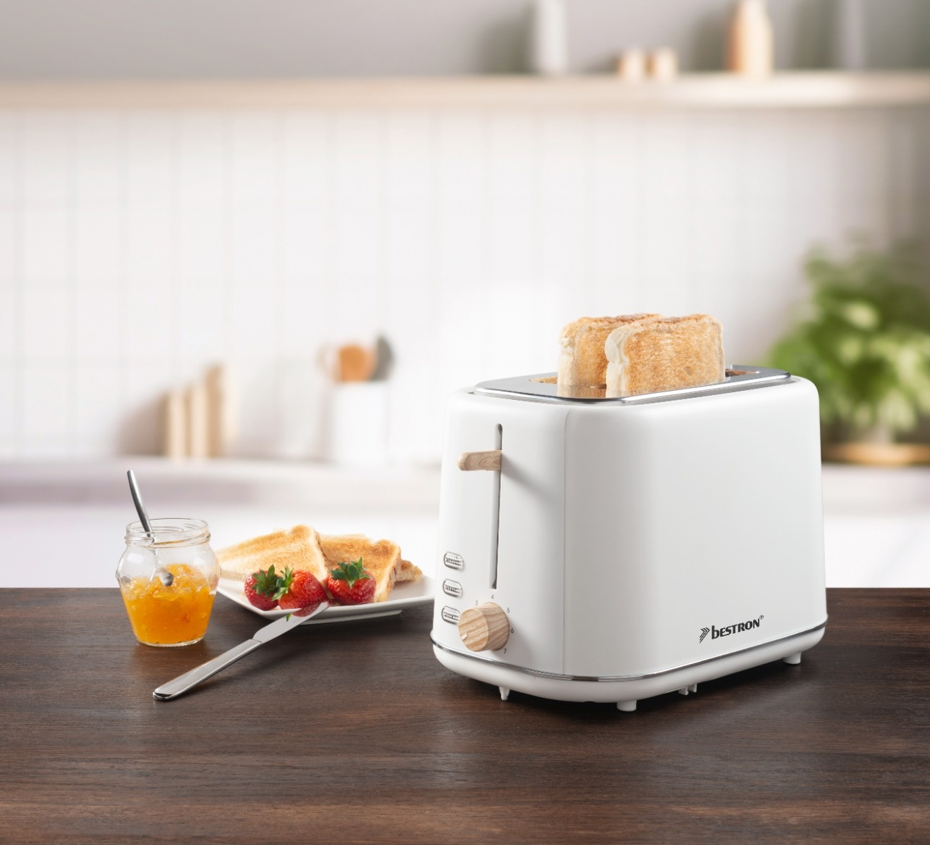 Ein weißer Toaster mit Toastbrot, Marmelade, Erdbeeren und einem Messer auf einem Holztisch. Ein Küchenhintergrund ist unscharf.