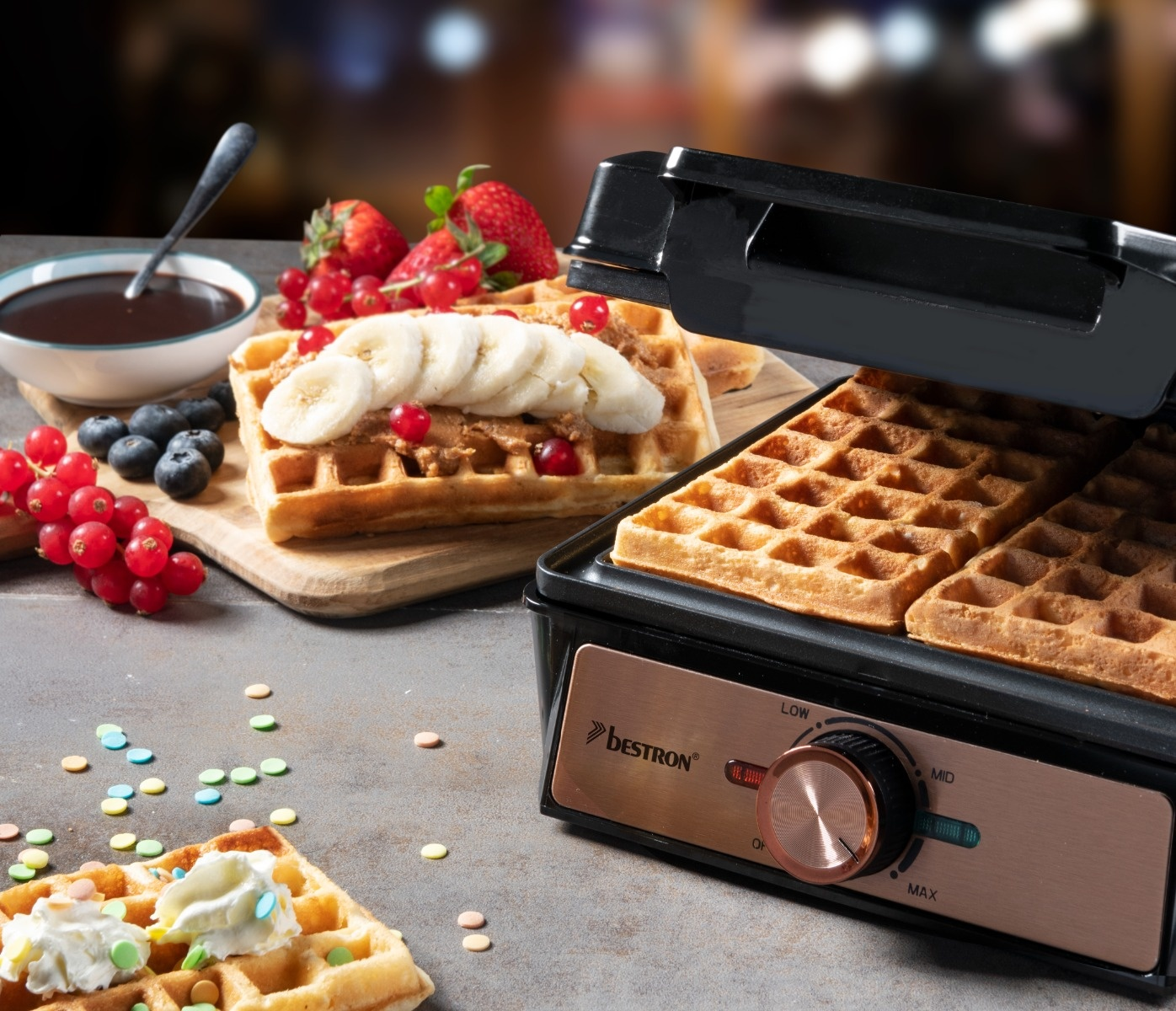 Waffeln mit Belag, Waffeleisen und Zutaten. Erdbeeren, Bananen, Blaubeeren und Johannisbeeren sind sichtbar.