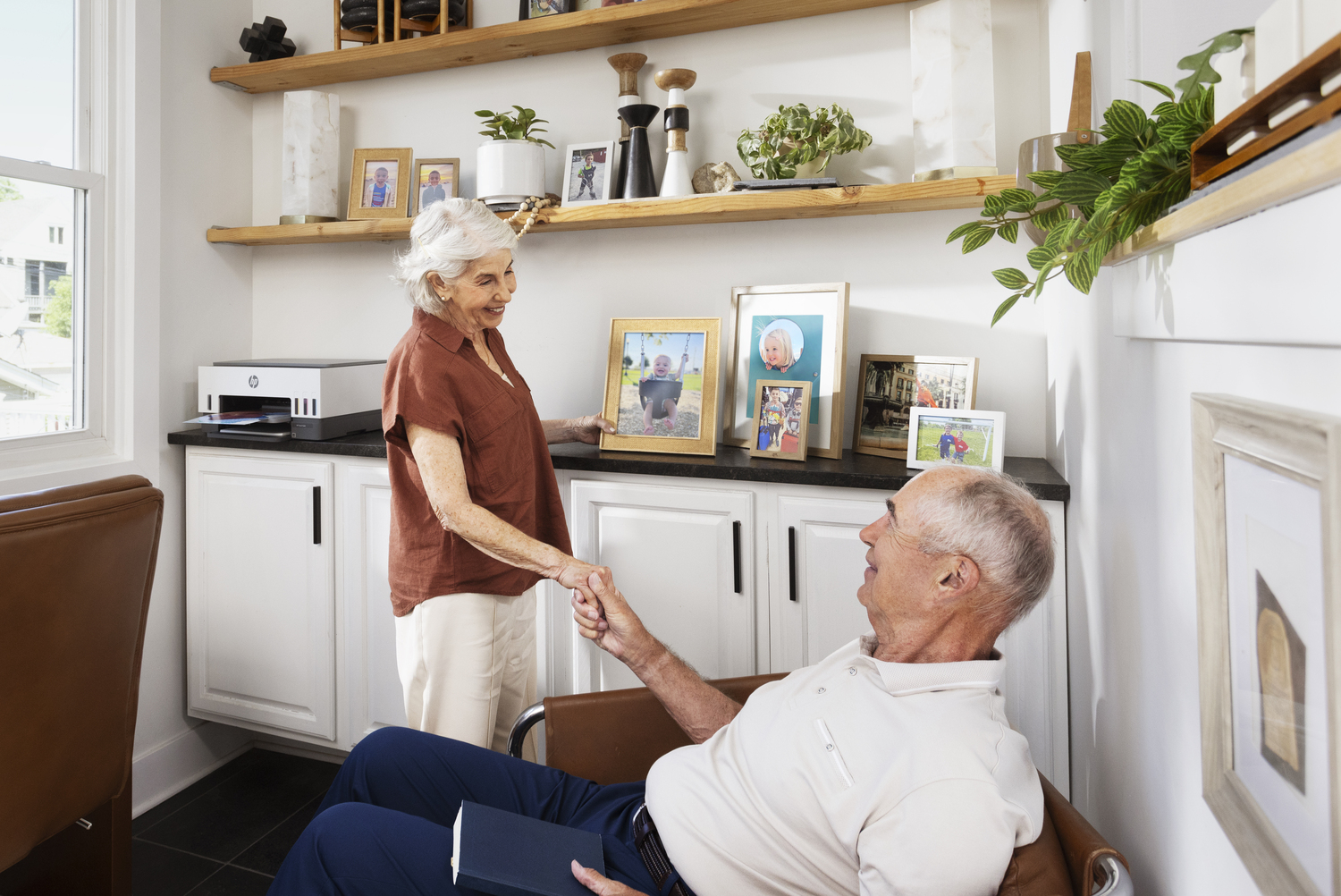 Una pareja de ancianos se da la mano. Una mujer con camisa marrón está de pie cerca de un hombre sentado en una silla marrón, sonriendo y mirándose.