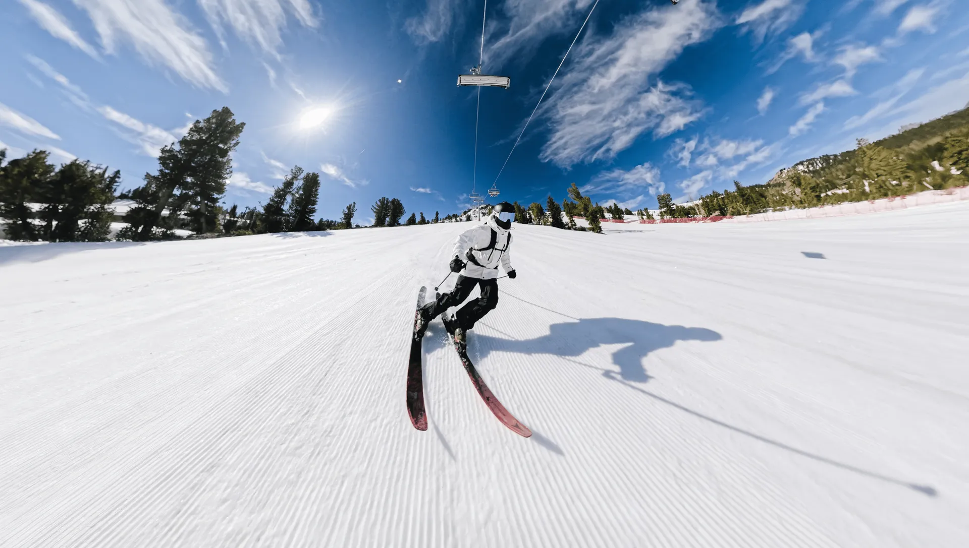 Skifahrer in weißer Jacke fährt einen schneebedeckten Hang hinunter. Blauer Himmel mit Wolken. Skilift im Hintergrund.
