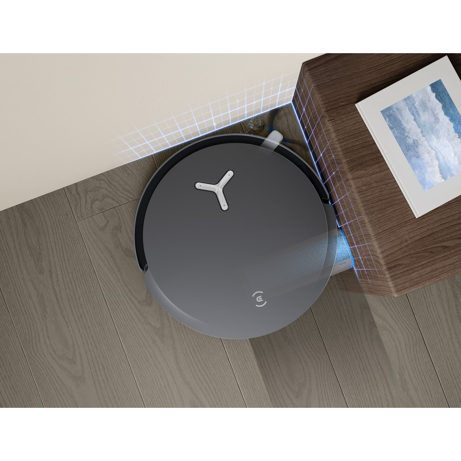 Un aspirateur robot gris est près d'une table en bois. Un cadre photo est sur la table. Le sol est en bois.