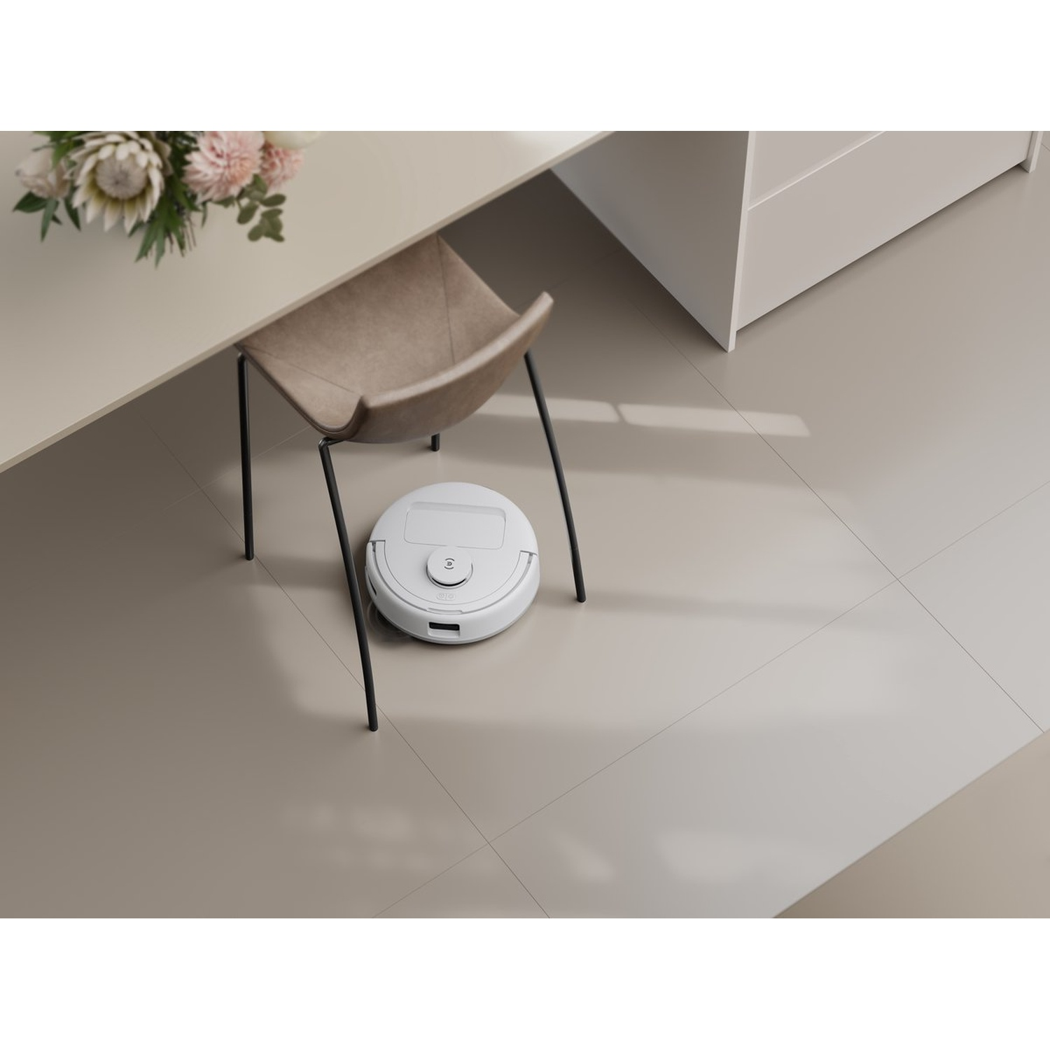 Een witte robotstofzuiger staat op een tegelvloer. Een stoel, tafel en bloemen staan op de achtergrond.