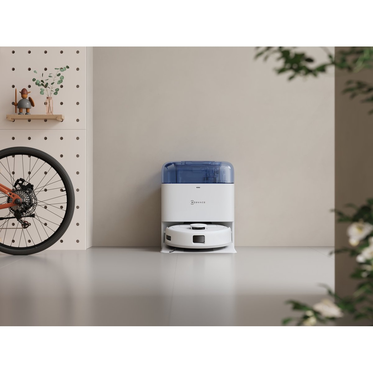 Een witte robotstofzuiger staat op een grijze vloer. Een fietswiel, plank met decoratie en een witte muur op de achtergrond.