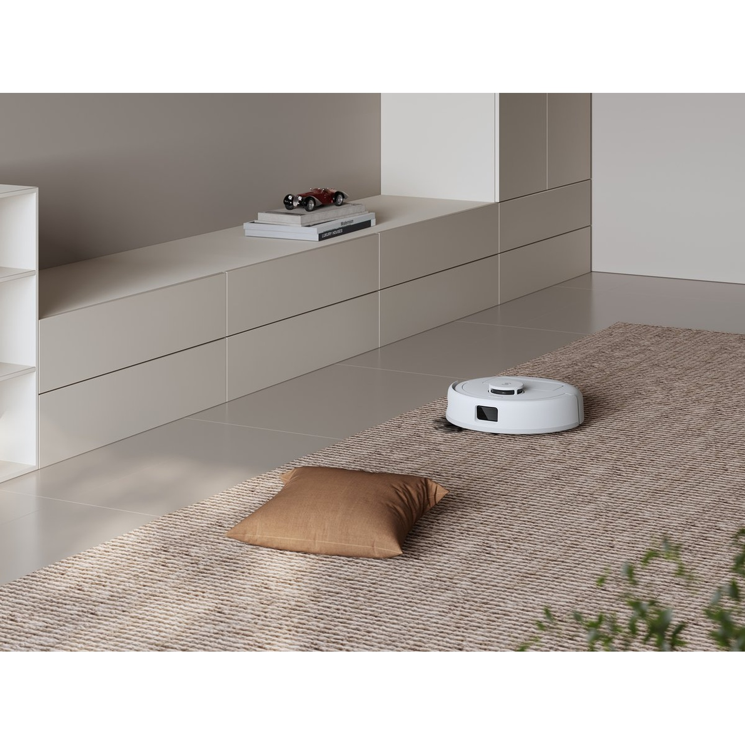 Een witte robotstofzuiger op een beige tapijt, een bruin kussen en een rode speelgoedauto op een plank.