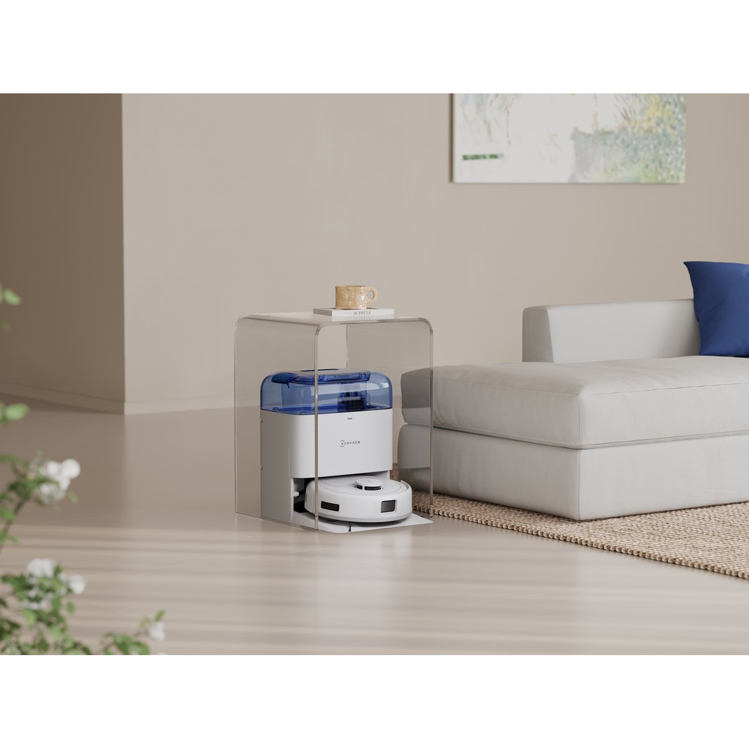 Een witte robotstofzuiger staat onder een doorzichtige tafel naast een witte bank met een blauw kussen. Er staat een kopje op de tafel.