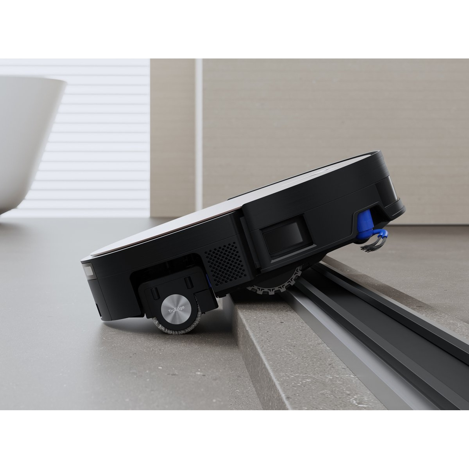 Un aspirateur robot noir sur une surface grise, s'approchant d'une voie encastrée. L'aspirateur a un dessus blanc et des accents bleus.