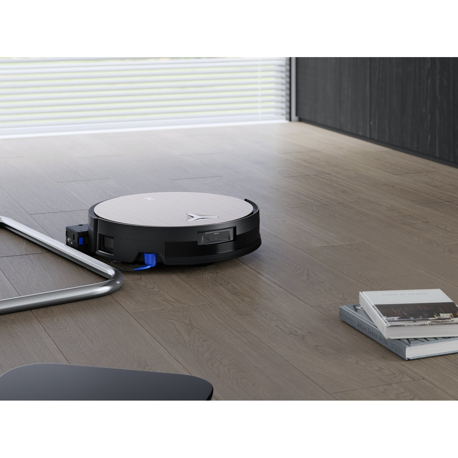 Un aspirateur robot sur un plancher en bois. Des livres et un objet métallique sont à proximité. Des stores sont visibles en arrière-plan.