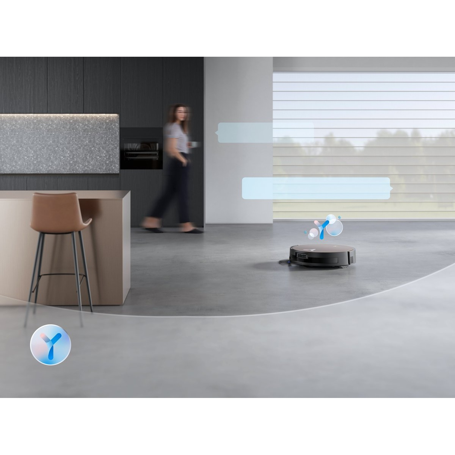 Un aspirateur robot sur un sol gris. Une personne floue marche en arrière-plan. Un tabouret et des meubles de cuisine à gauche.