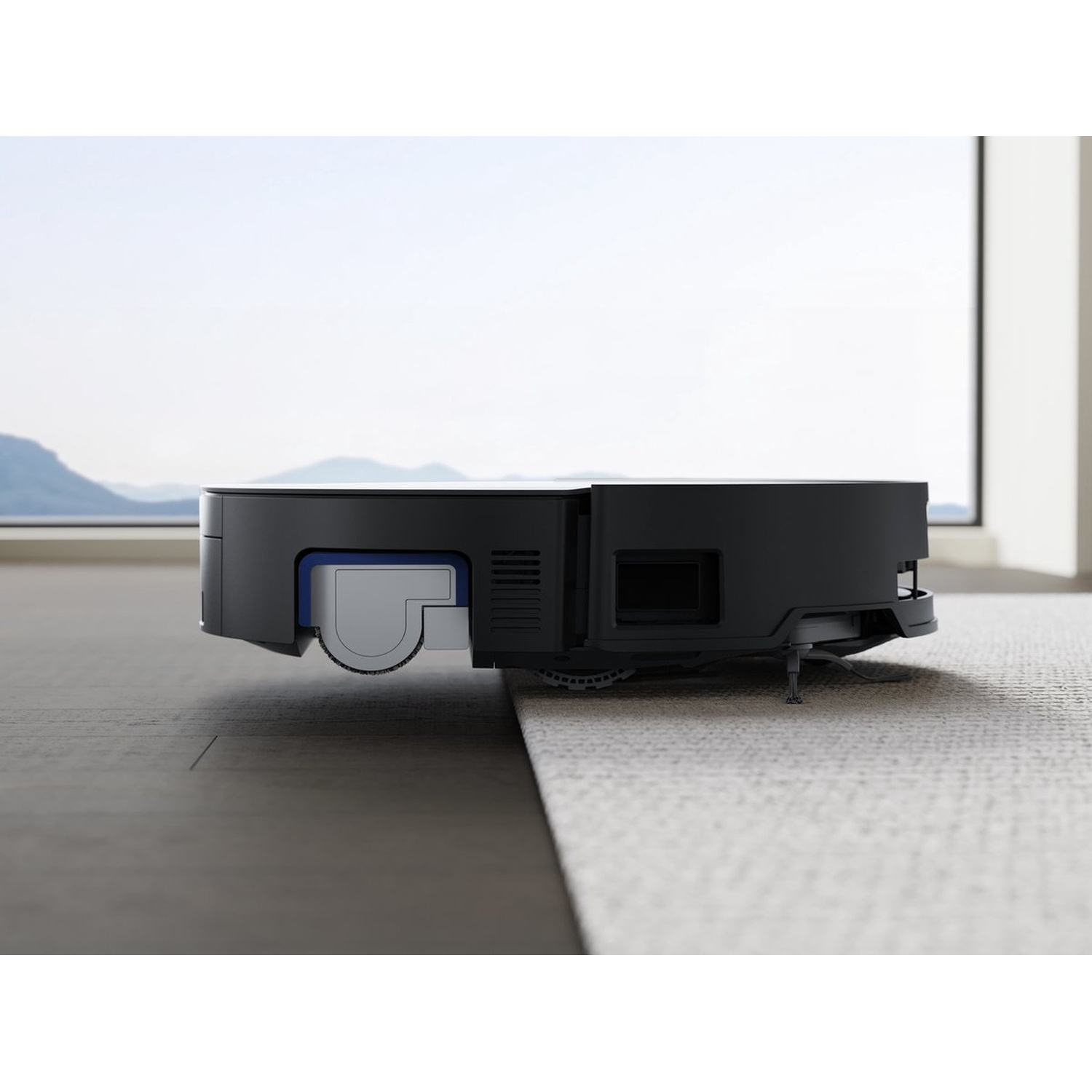 Un aspirateur robot noir sur un tapis clair. L'arrière-plan montre une fenêtre avec vue sur le ciel et un mur.