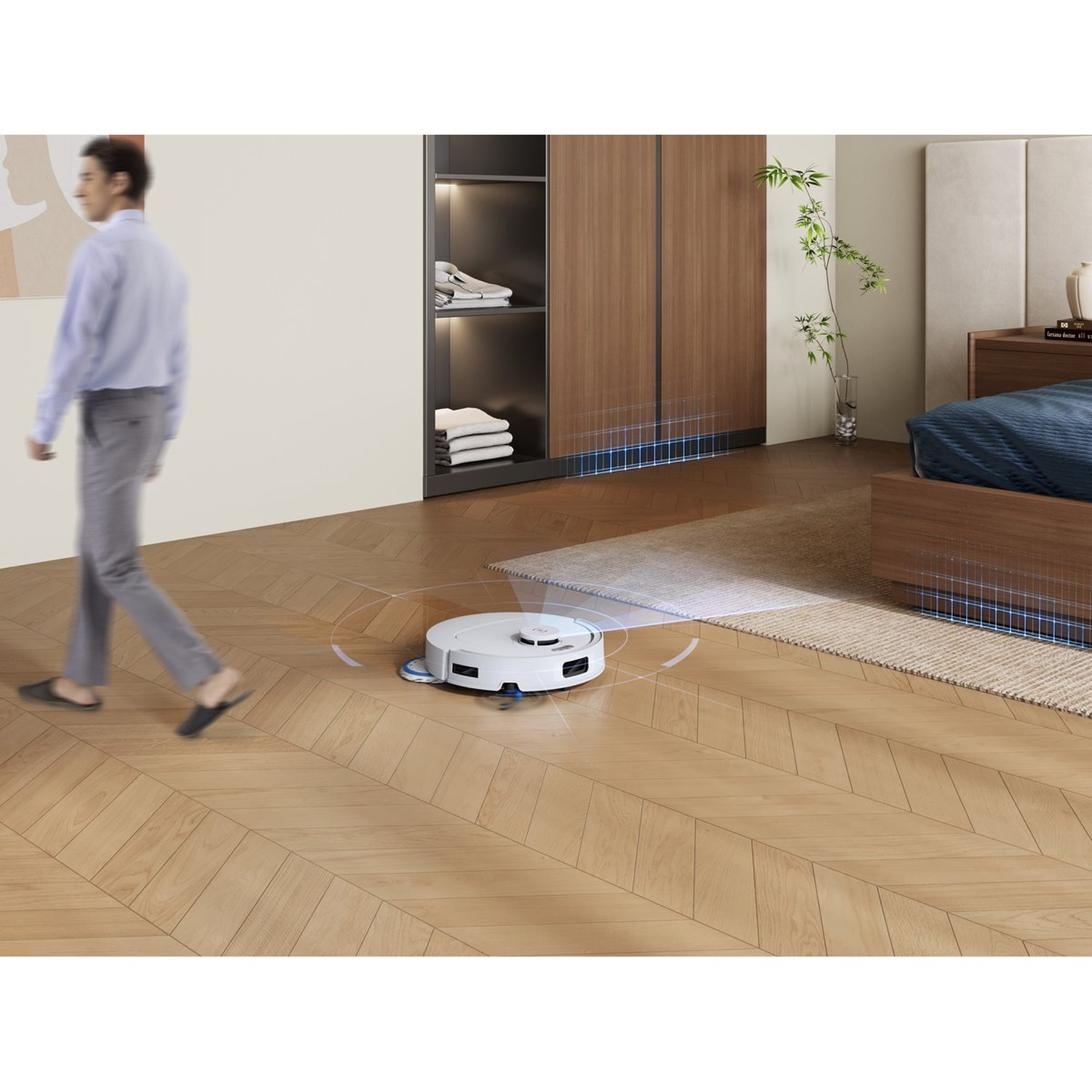 Een man loopt langs een robotstofzuiger in een slaapkamer. De robot reinigt een houten vloer. Een bed en een kast staan op de achtergrond.