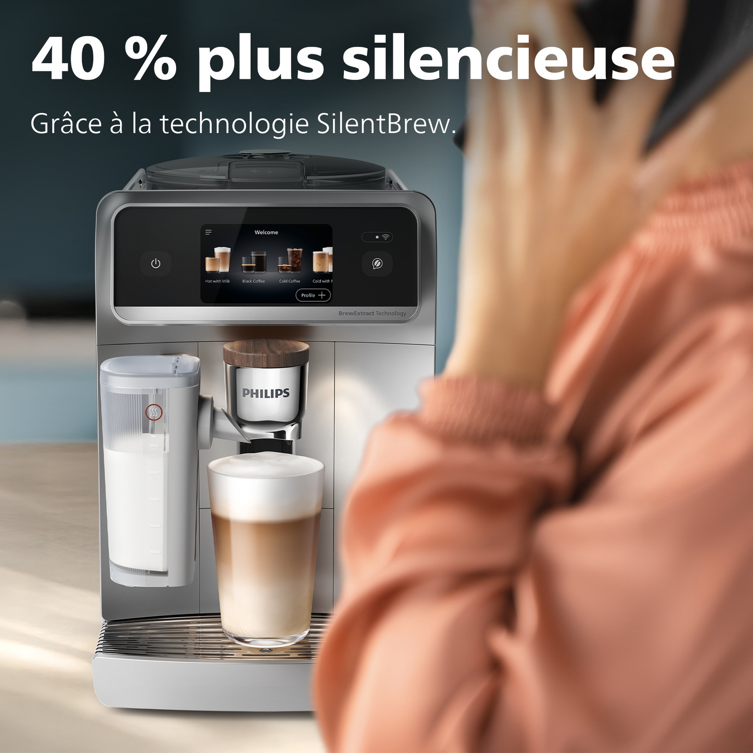 Une machine à café avec un écran affiche des options de café. Une personne est floue en arrière-plan. La machine a un verre de café.