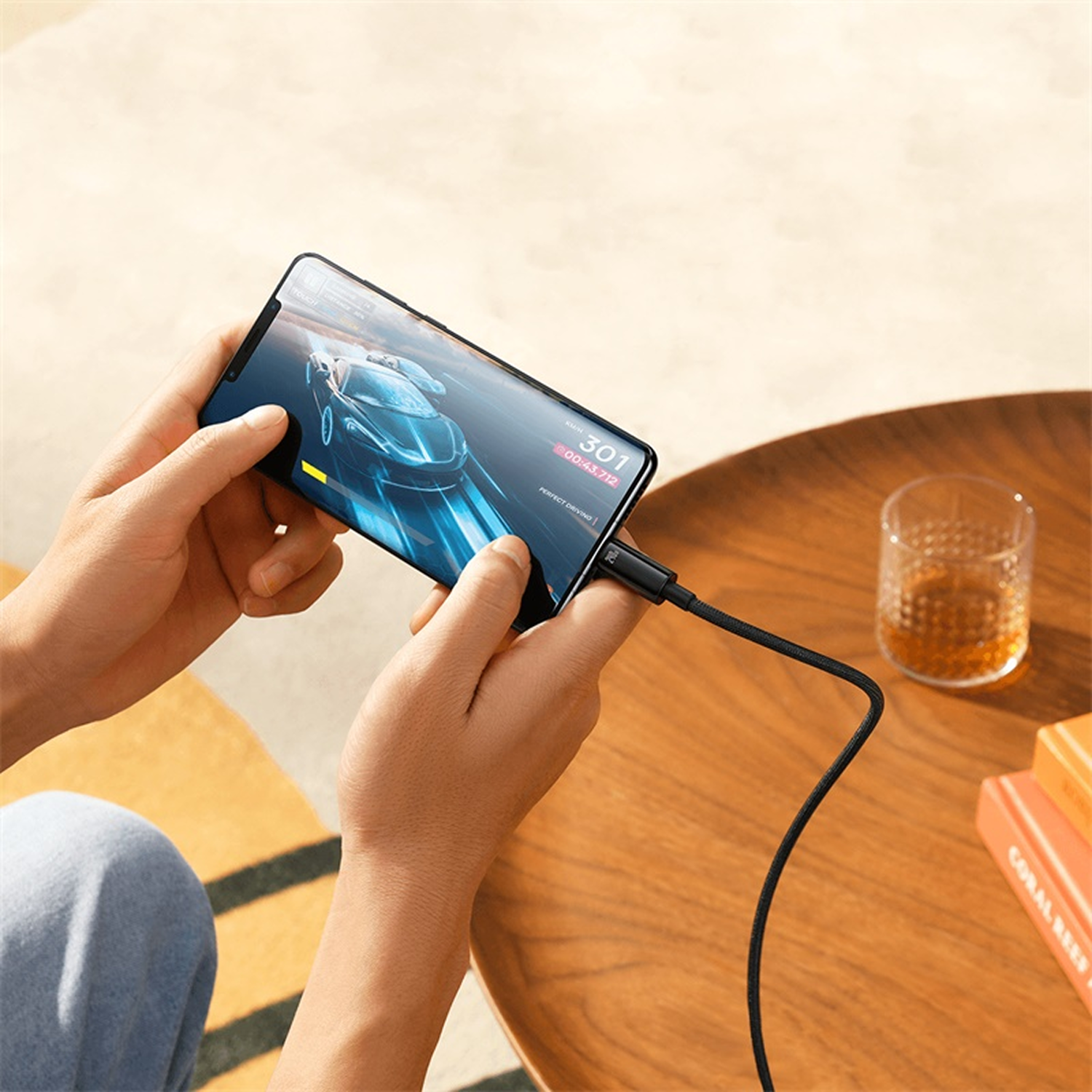 Manos sosteniendo un teléfono con un juego de carreras de autos, conectado a un cable, sobre una mesa de madera con un vaso y libros.