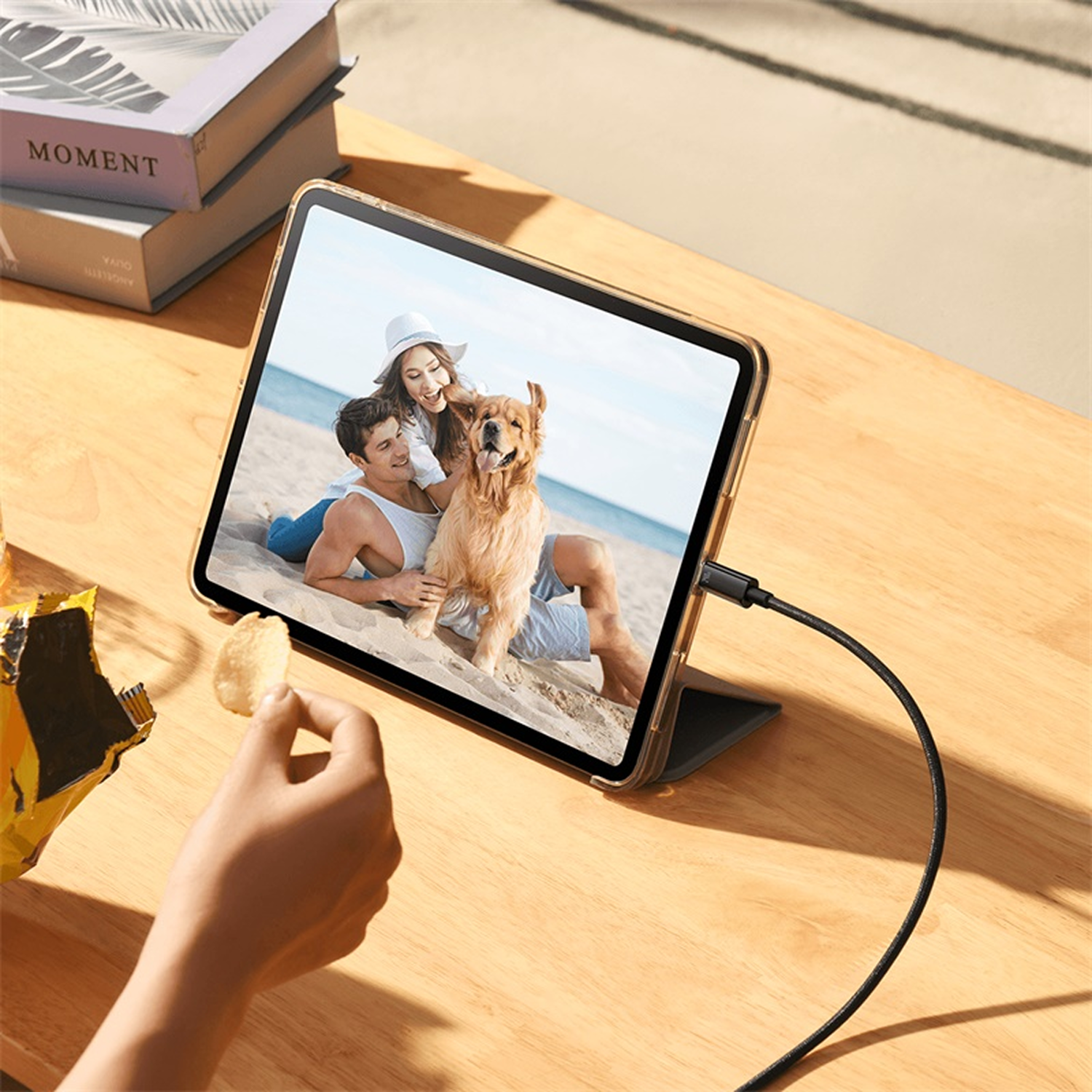 Tableta muestra foto de playa de pareja con perro. Mano sosteniendo chip. Libros a la izquierda. Cable conectado.