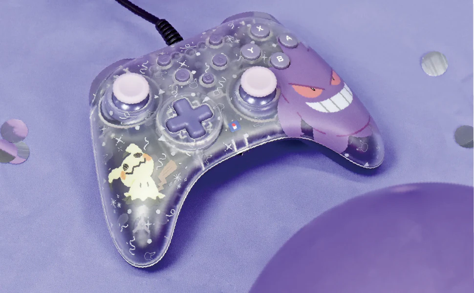 Ein klarer Pokemon-Controller mit lila Knöpfen, Gengar- und Mimikyu-Designs, auf einer lila Oberfläche mit Konfetti und einem Ballon.