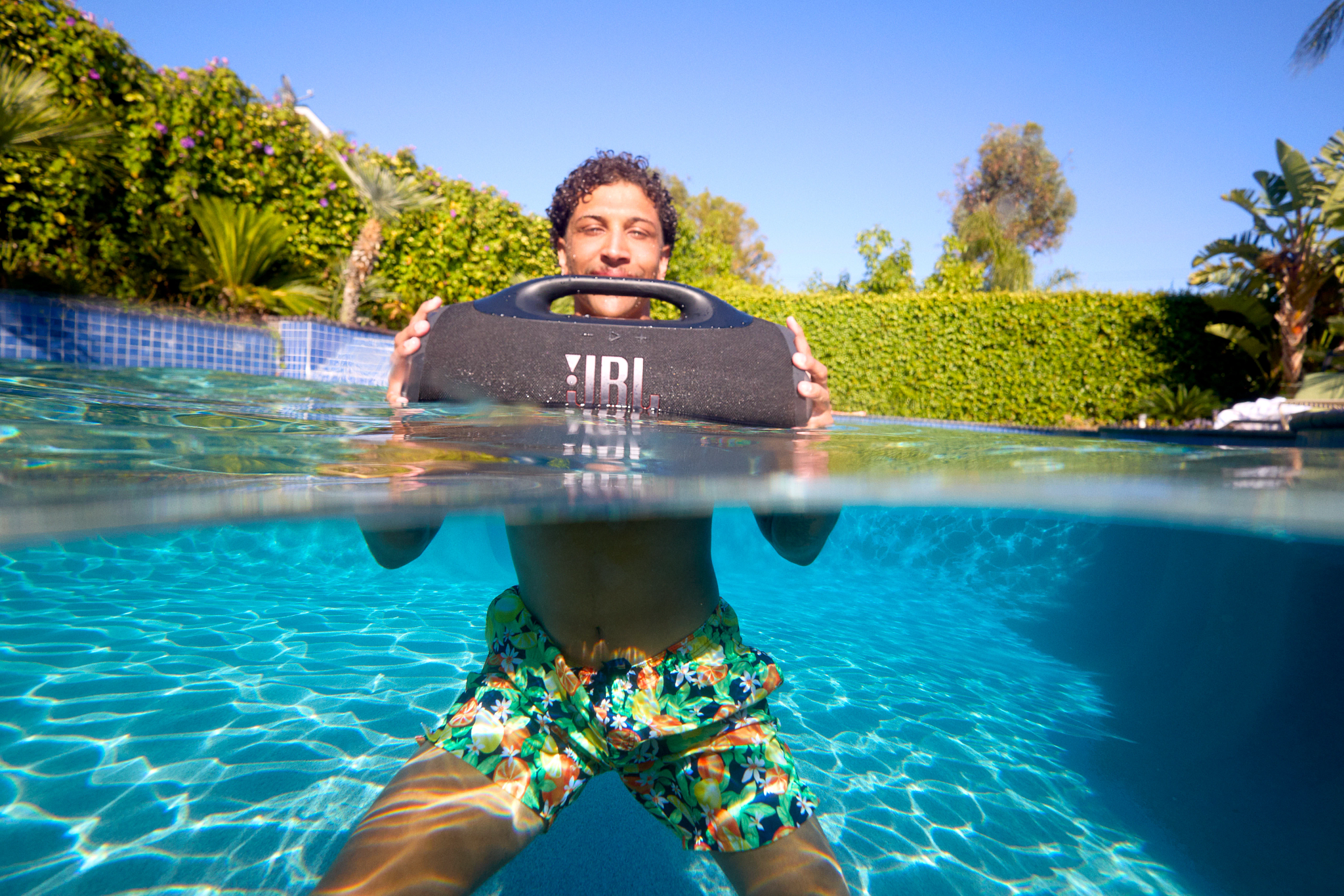Une personne dans une piscine tient un haut-parleur JBL. Elle porte un short de bain fleuri. Le fond montre une haie et un ciel bleu clair.