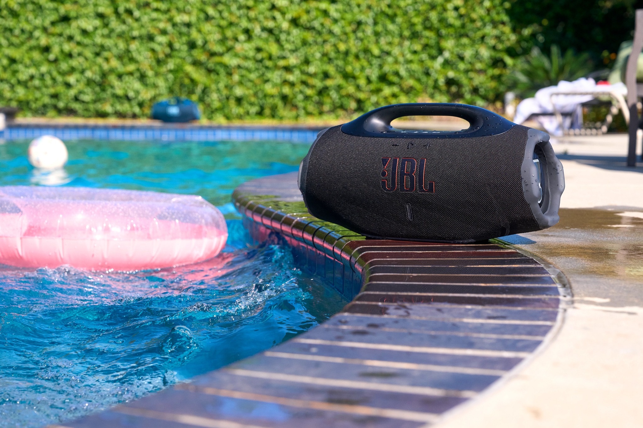 Une enceinte JBL noire au bord de la piscine. Un gonflable rose et une balle blanche flottent dans l'eau bleue. Fond de haie verte.