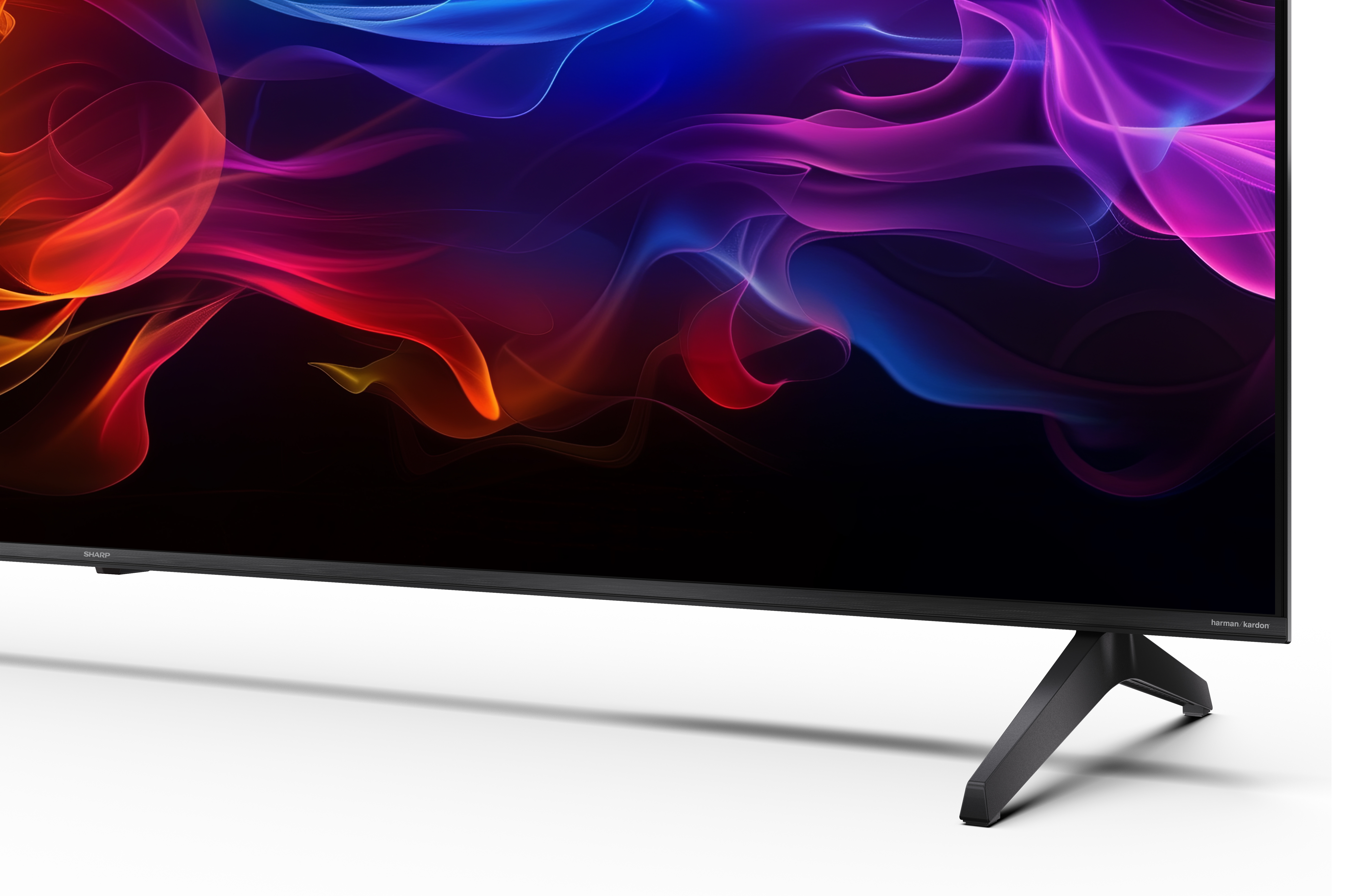 Egy Sharp TV színes absztrakt mintákat jelenít meg. Fekete lábai vannak, alján a "harman/kardon" felirattal.
