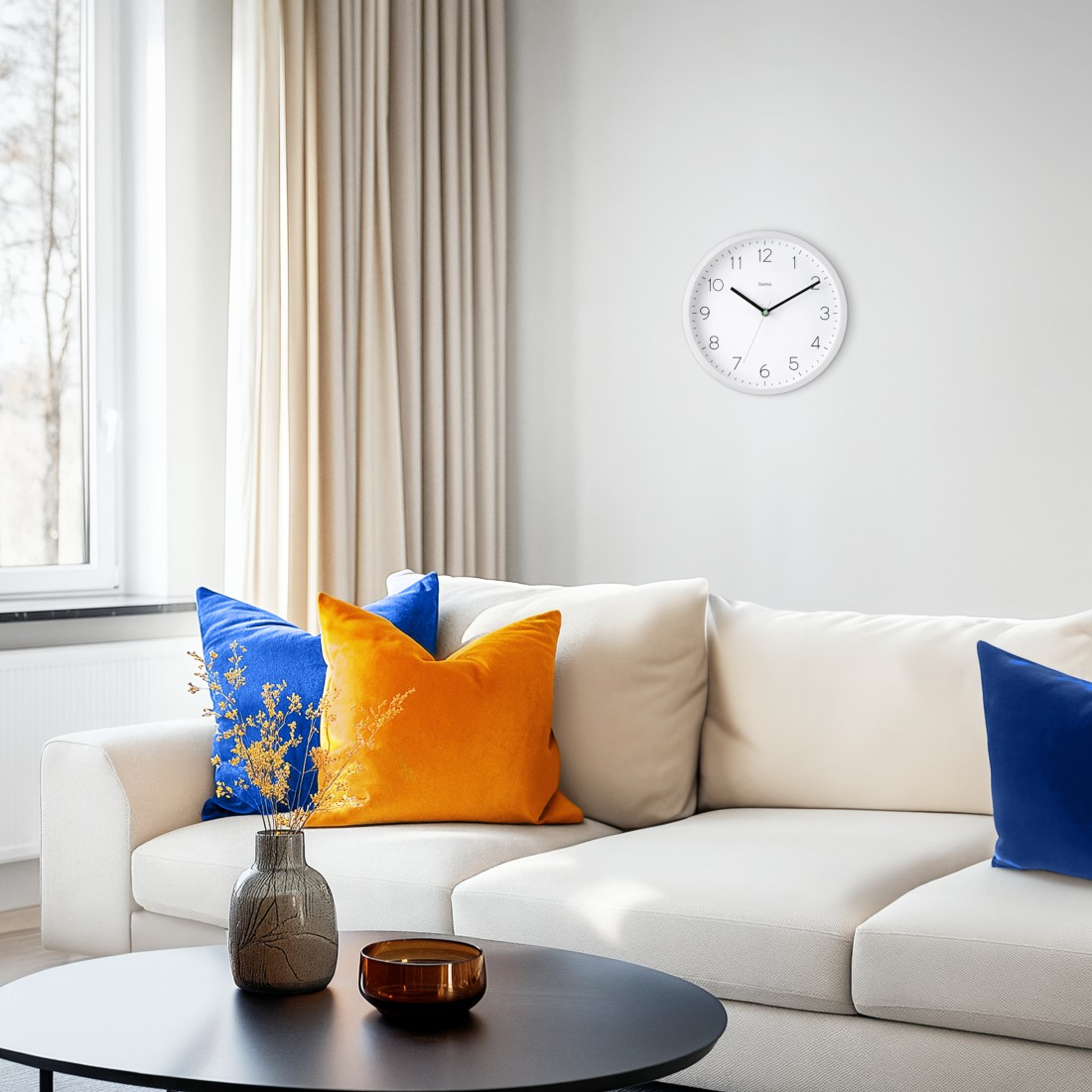 Wohnzimmer mit weißem Sofa, bunten Kissen, Uhr an der Wand, Vase und Tisch. Fenster und Vorhänge im Hintergrund.