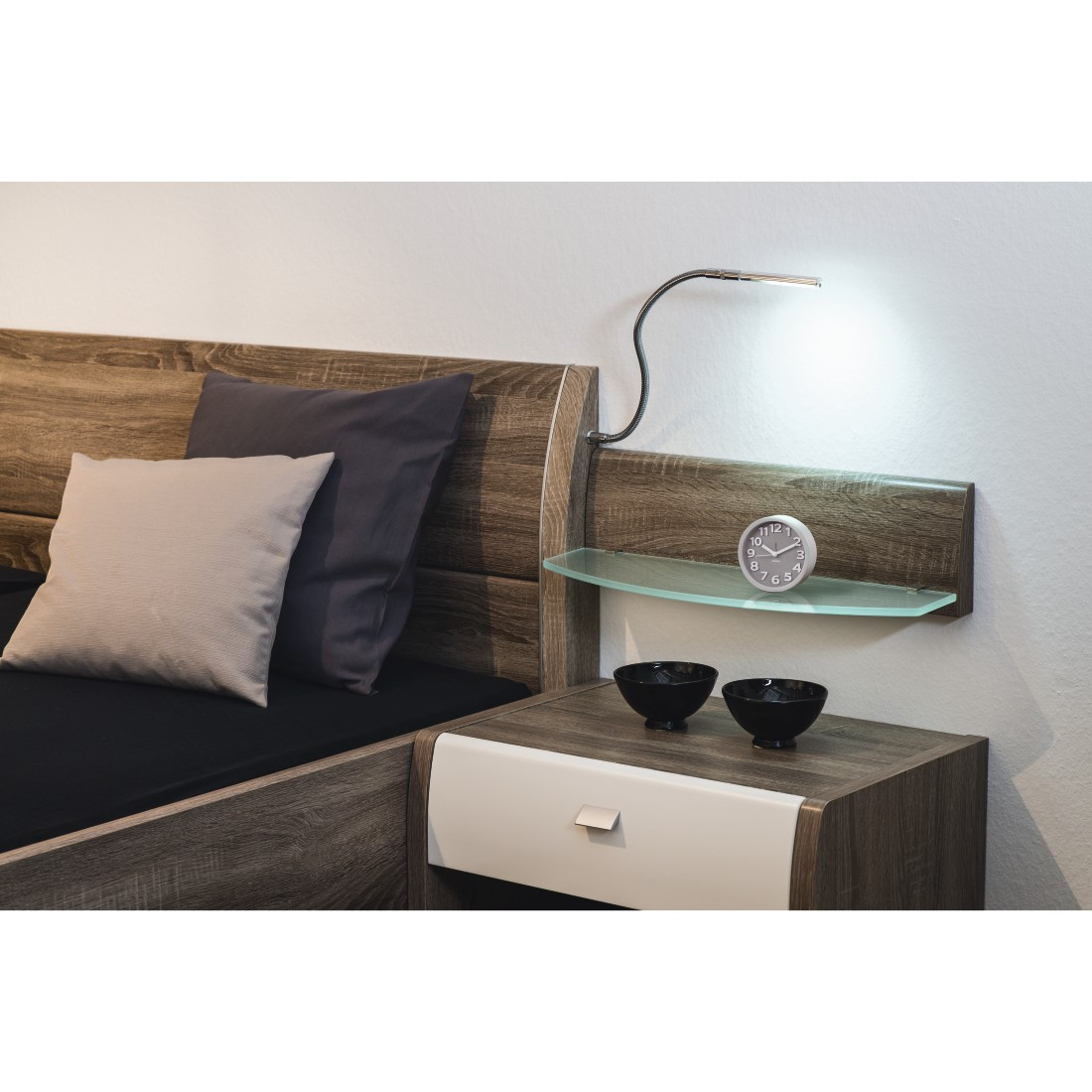 Schlafzimmer-Szene. Bett mit Kissen, ein Nachttisch mit Uhr und zwei Schalen. Eine Lampe scheint auf das Regal.