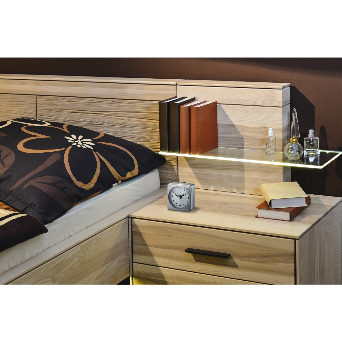 Schlafzimmer mit Holzbett, Nachttisch und Regal. Bücher, Uhr und Parfums sind sichtbar. Das Bett hat ein gemustertes Kissen.