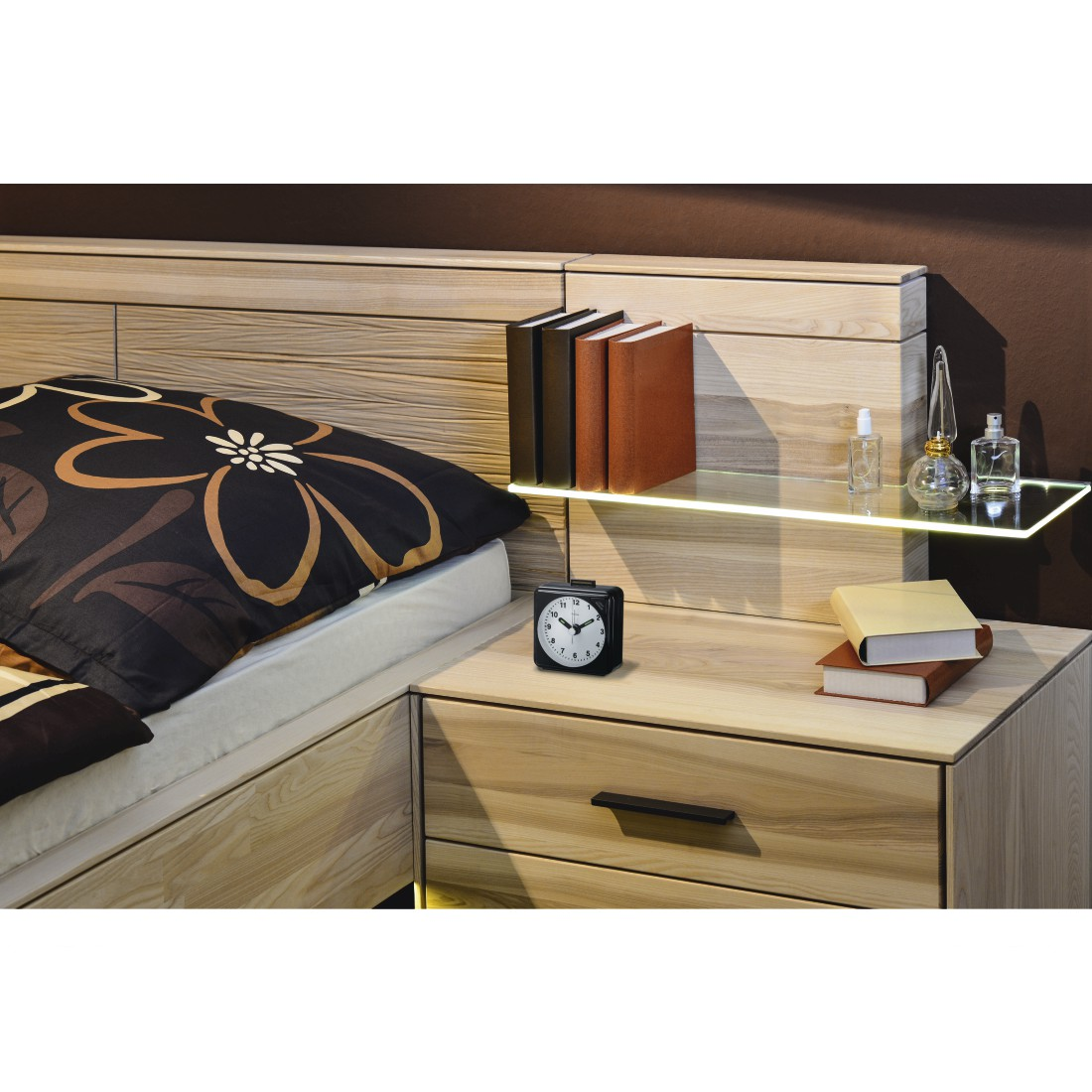 Ein Schlafzimmer mit Bett, Nachttisch, Büchern und Wecker. Bücher stehen auf einem Glasregal. Das Bett hat eine gemusterte Tagesdecke.