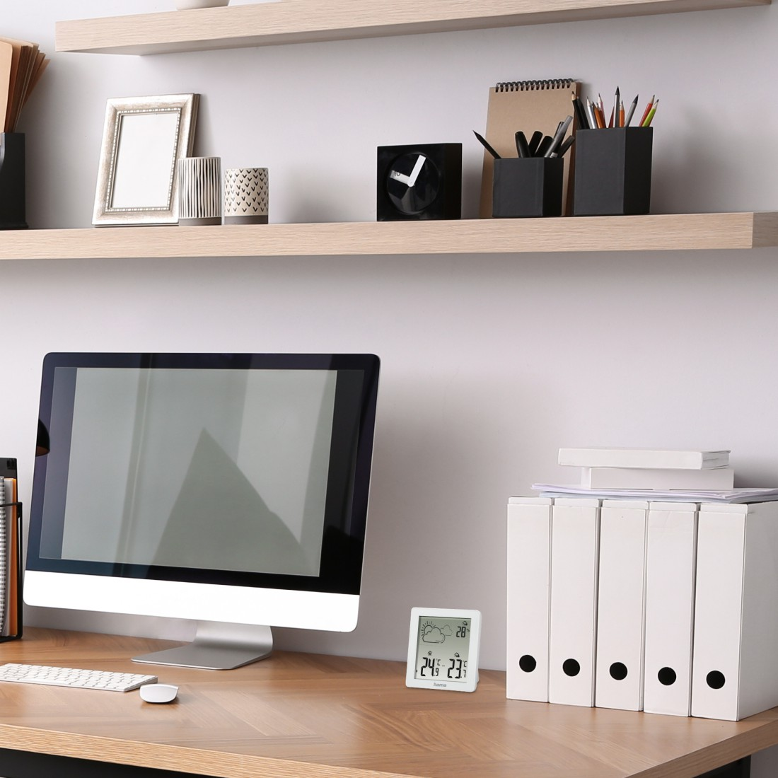 Schreibtisch mit Computer, Akten und Wetterstation. Regale oben halten Dekorationsgegenstände. Der Schreibtisch ist holzmaseriert.
