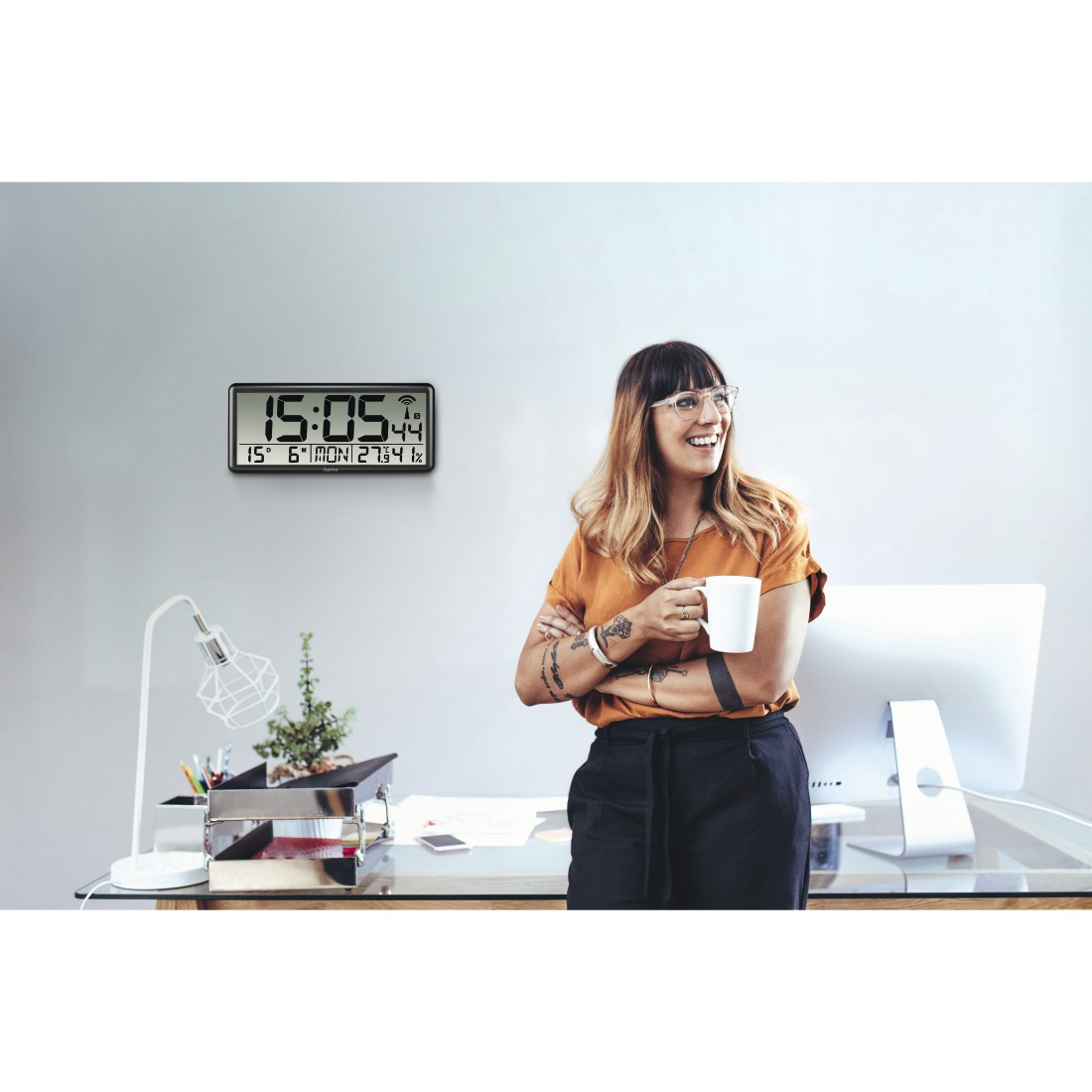 Frau in orangefarbenem Hemd hält eine Tasse und lächelt in der Nähe eines Schreibtisches. Eine Uhr hängt an der Wand. Der Schreibtisch hat einen Computer, eine Lampe und Gegenstände.