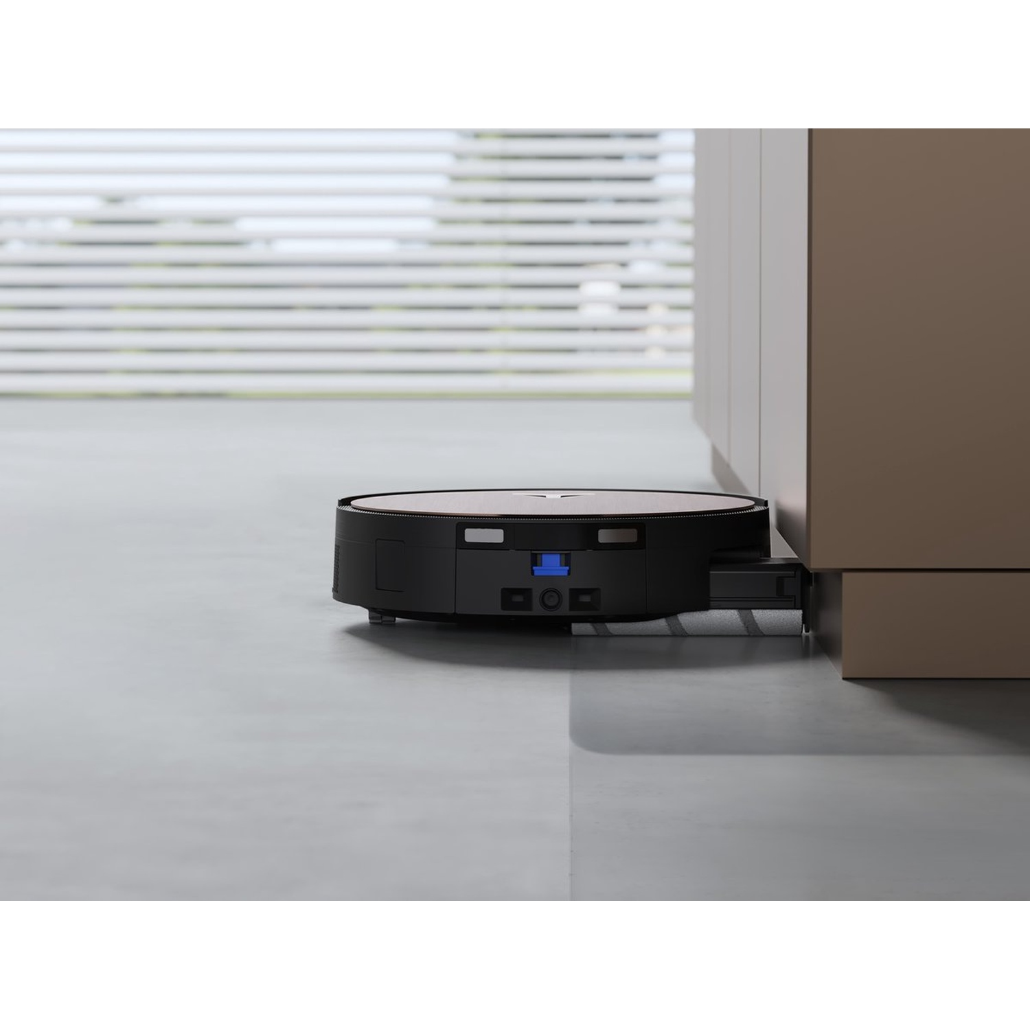 A black and silver robot vacuum cleaner is cleaning the floor. It is next to a cabinet. Blinds are visible in the background.