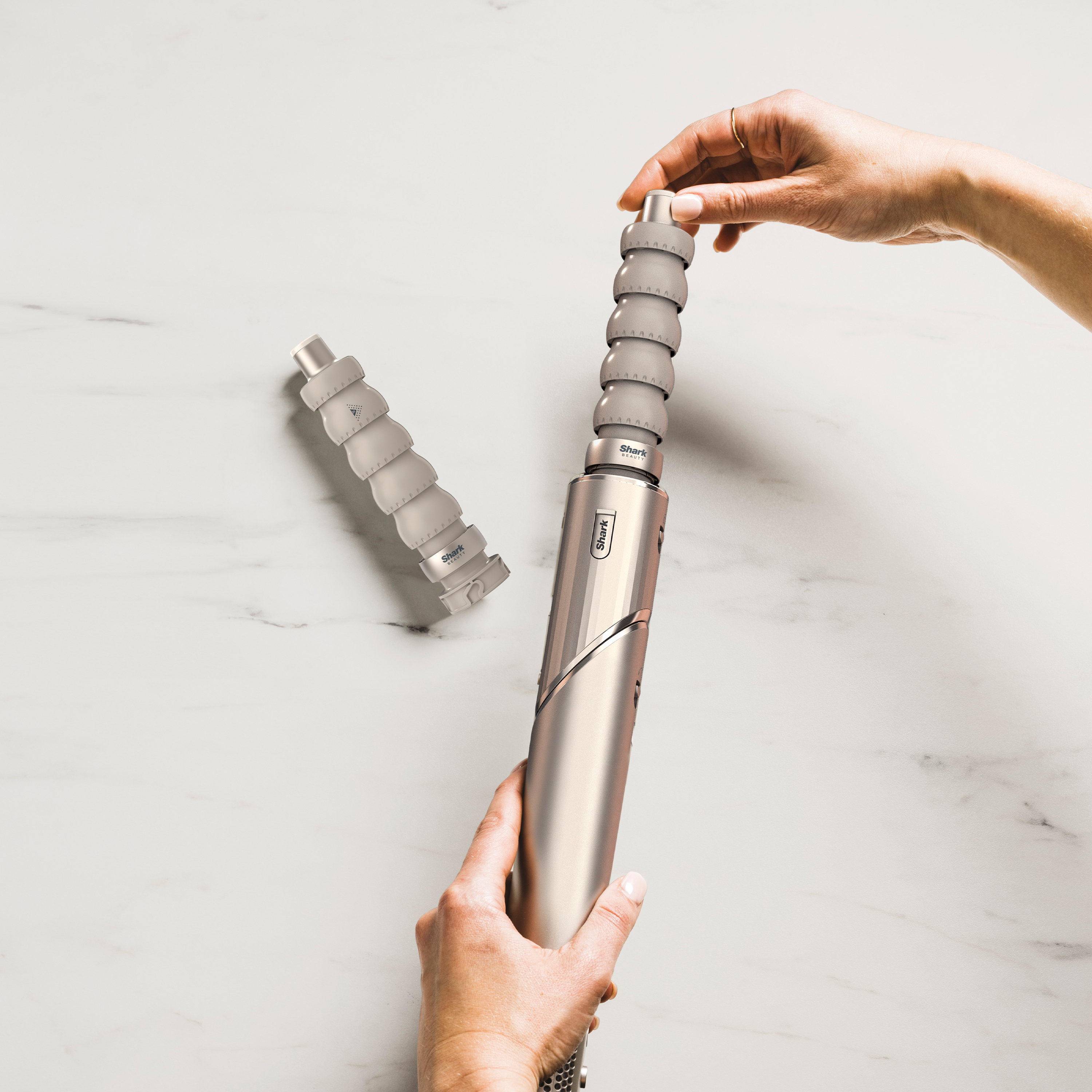 Ręce składające urządzenie Shark Beauty na marmurowej powierzchni. Komponenty są srebrne, szare i białe.