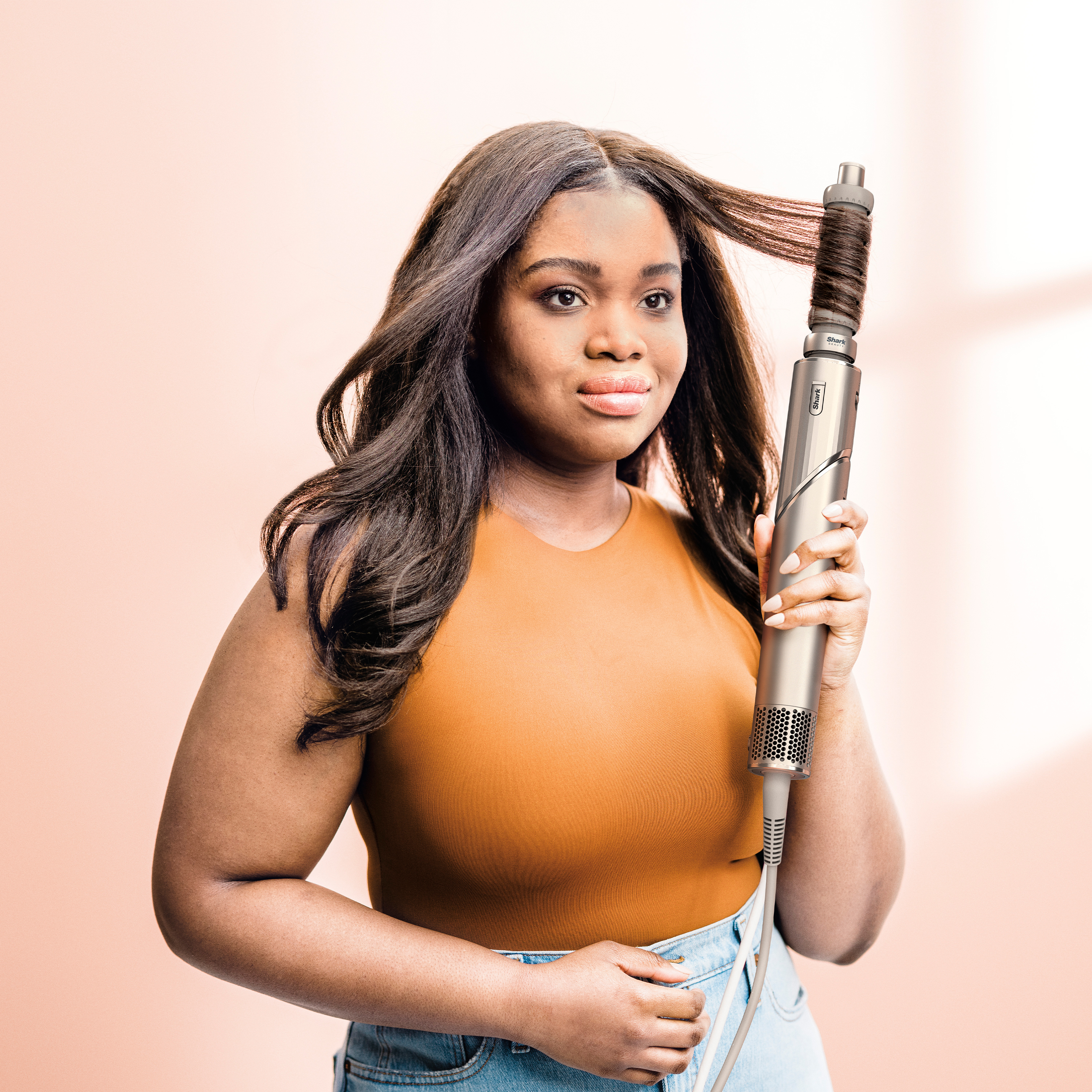 Kobieta trzyma prostownicę do włosów. Ma na sobie top w kolorze brązowym i dżinsy, z długimi ciemnymi włosami. Tło jest brzoskwiniowe.