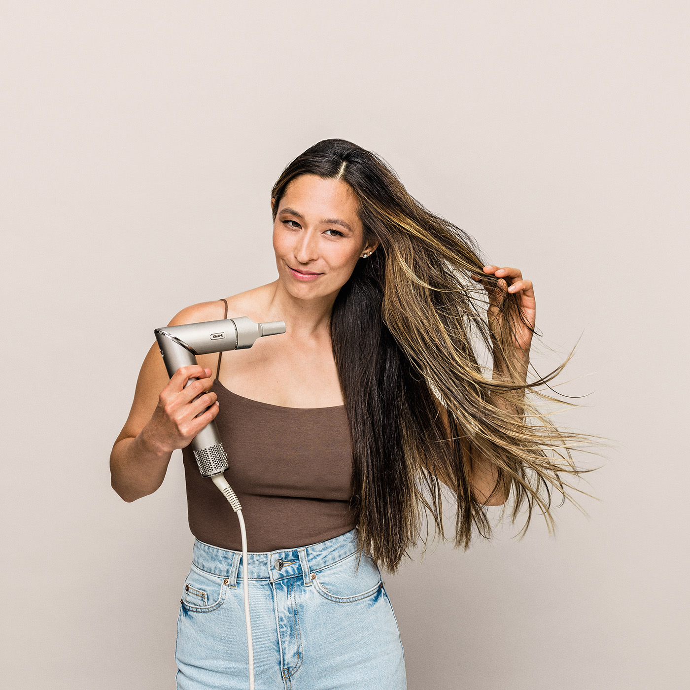 Kobieta trzyma suszarkę do włosów, dotyka włosów. Ma na sobie brązowy top i dżinsy. Tło beżowe.