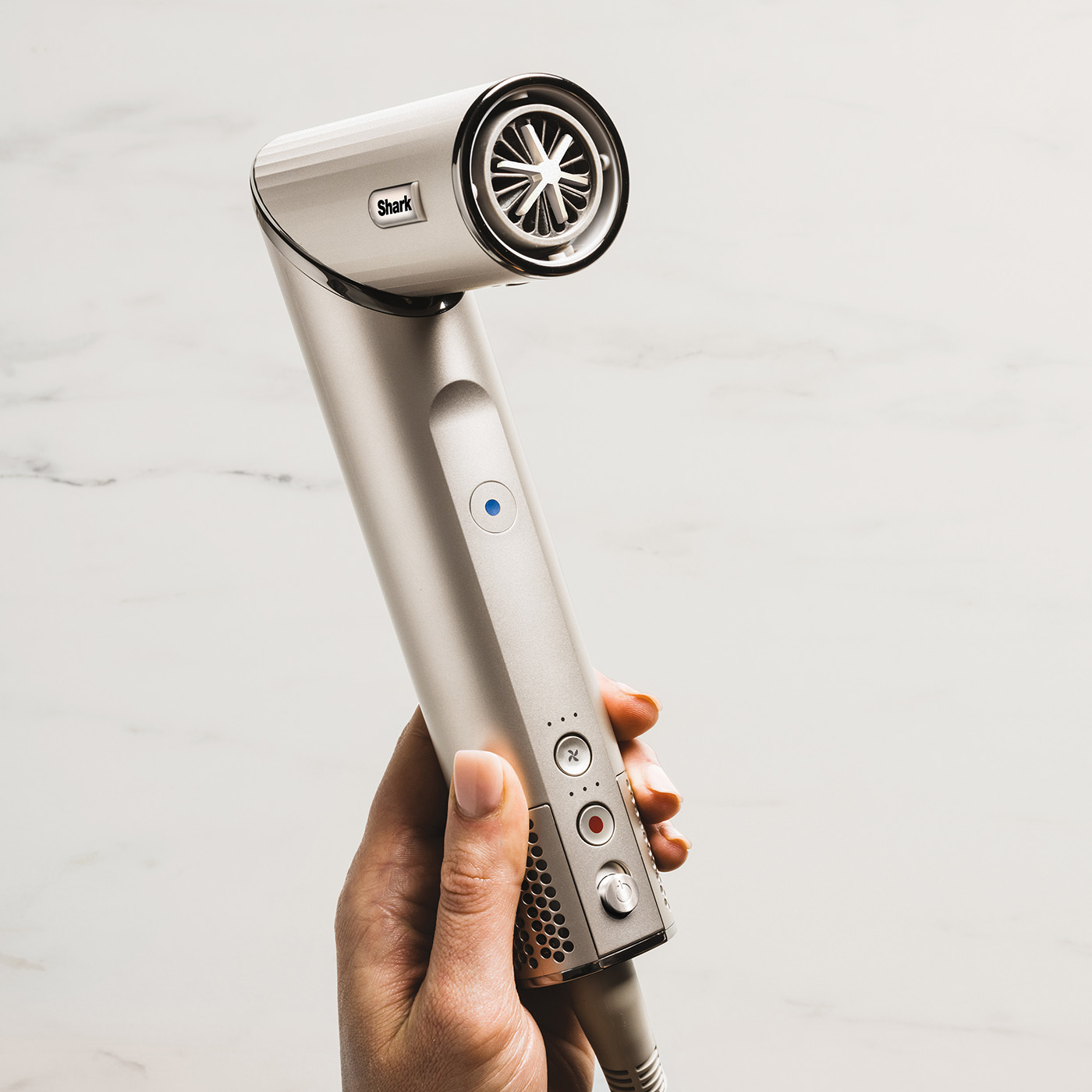 Dłoń trzyma srebrną suszarkę do włosów Shark. Suszarka ma dyszę, przyciski i przewód zasilający. Tło to biały marmur.