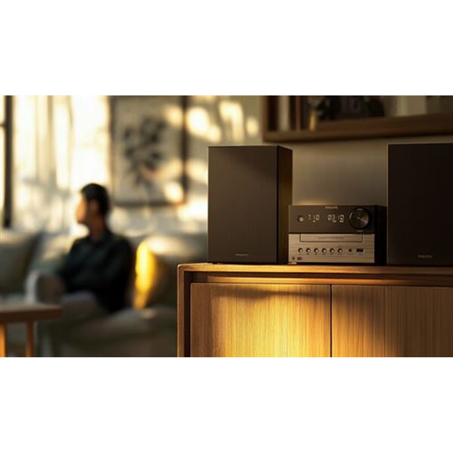 A stereo system sits on a wooden cabinet. A person sits on a couch in the background. Soft lighting illuminates the room.