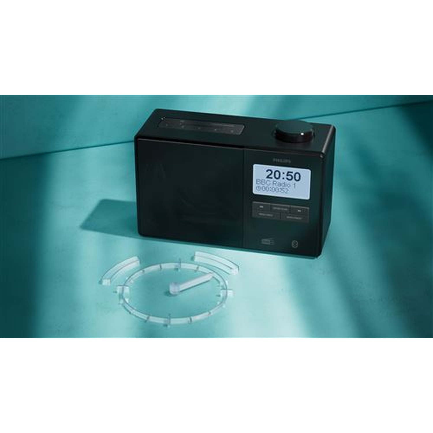 Black Philips radio displaying time and station. A transparent clock-like object sits in front. Turquoise background.