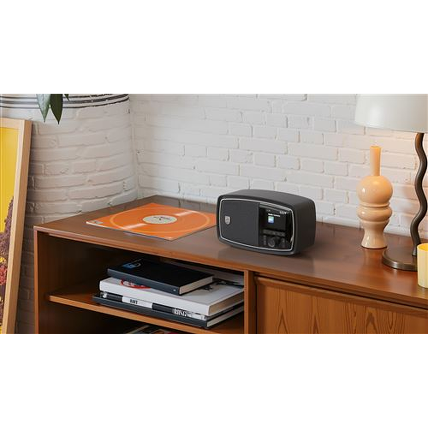 A retro radio sits on a wooden cabinet, with a vinyl record, books, and a lamp. White brick wall in the background.