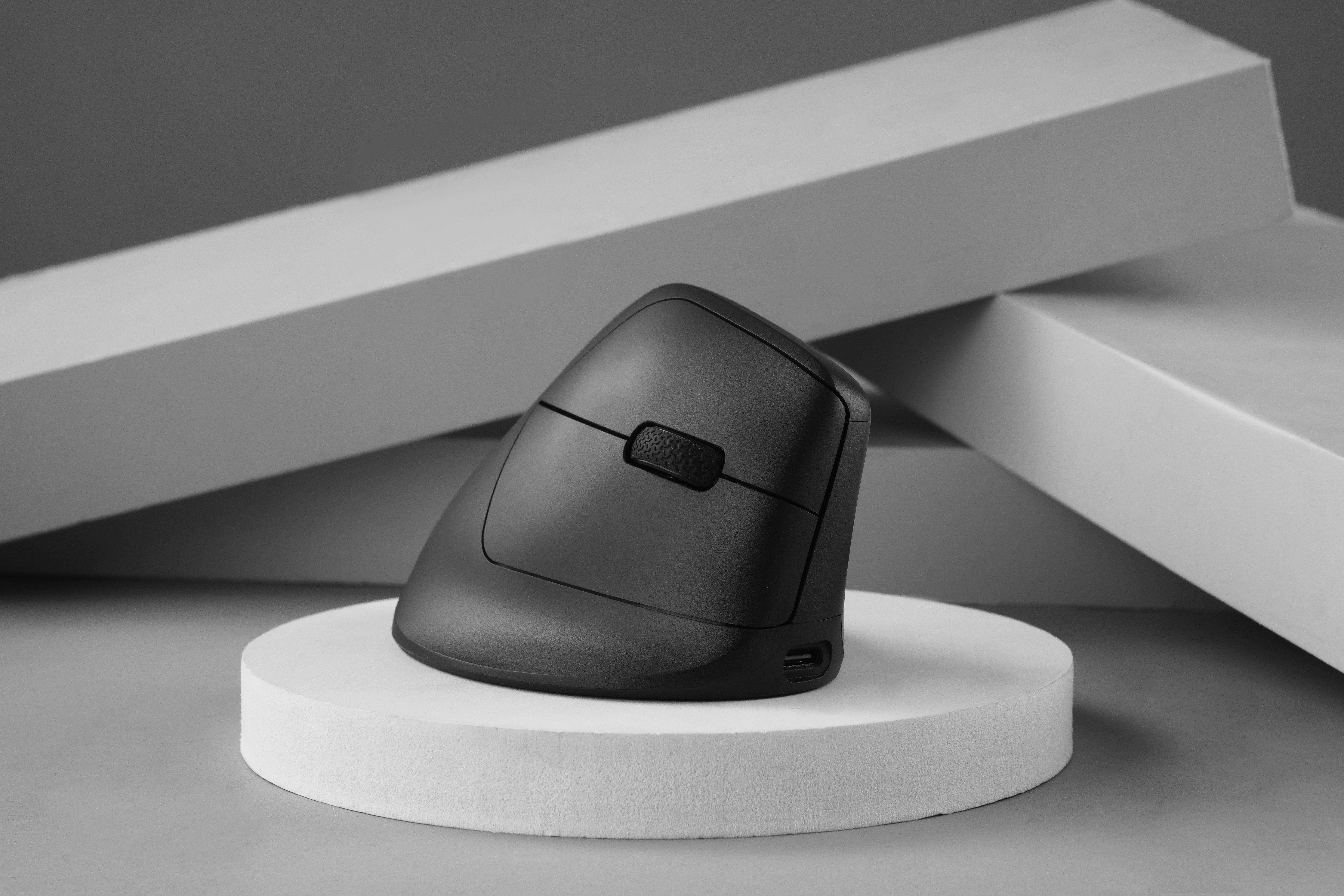 Schwarze ergonomische Maus auf einem weißen, kreisförmigen Ständer. Im Hintergrund weiße geometrische Formen.