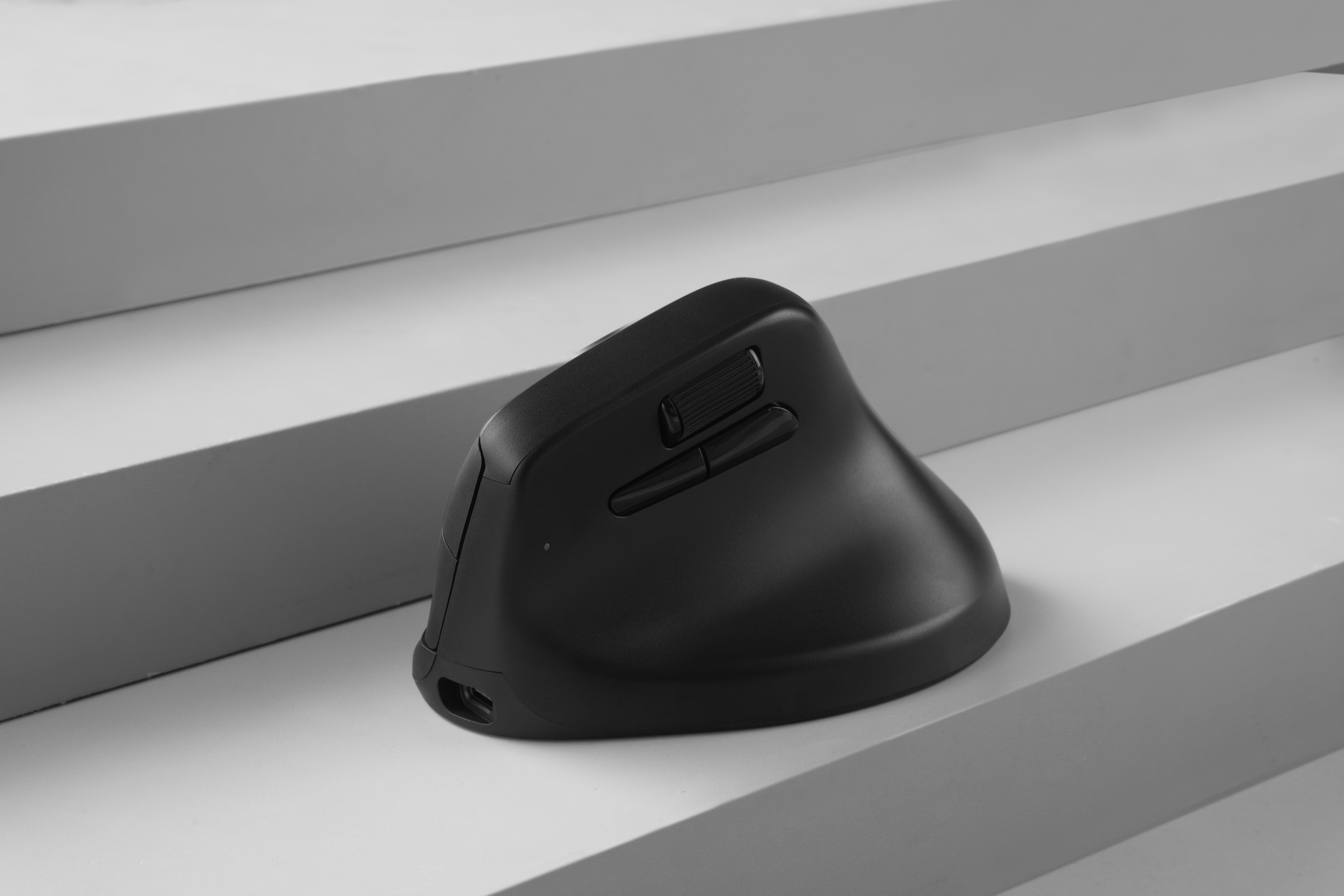 Eine schwarze ergonomische Computermaus liegt auf einer weißen, abgestuften Oberfläche. Die Maus hat Tasten und ein Scrollrad.