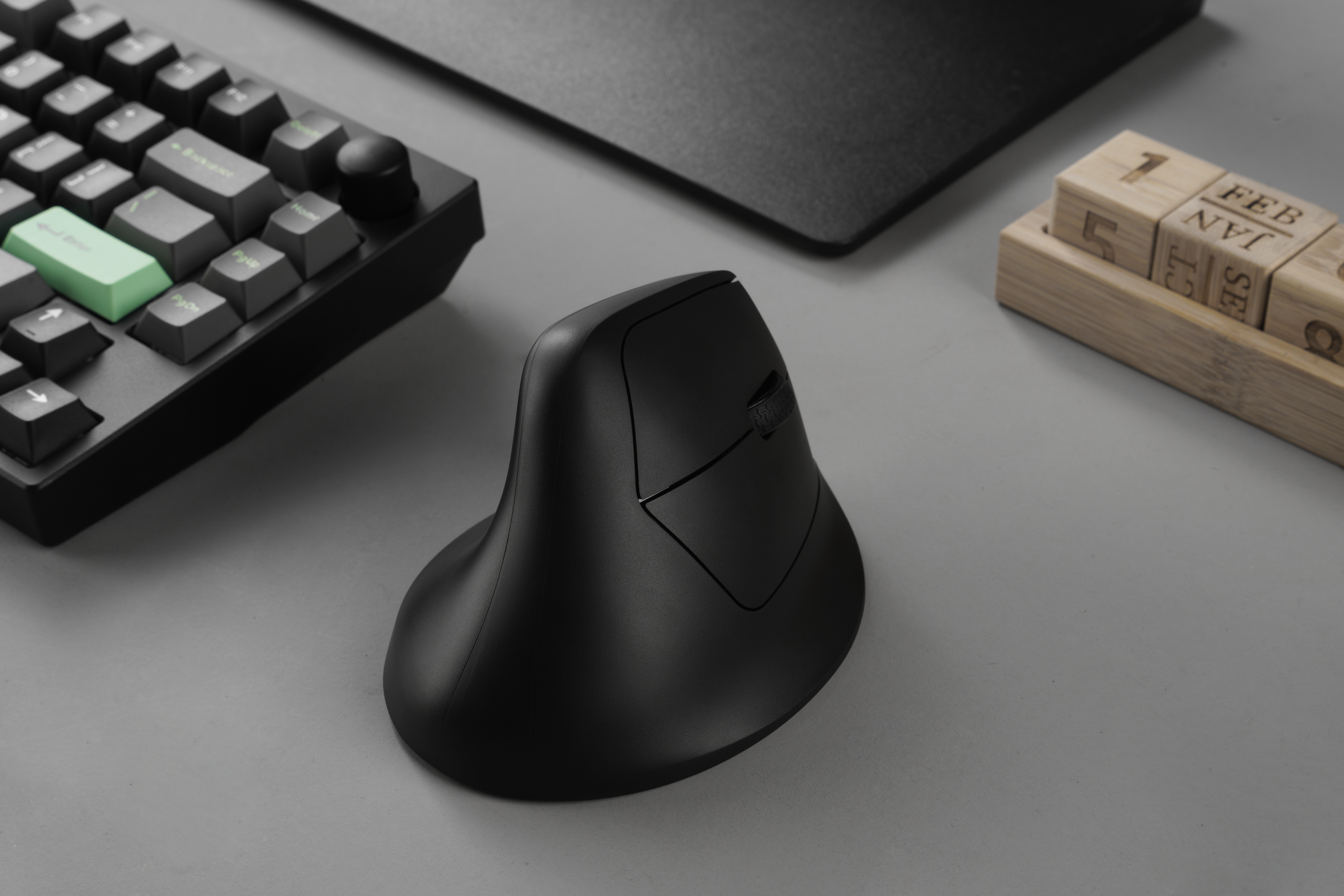 Schwarze ergonomische Maus, Tastatur und Holzkalender auf grauer Oberfläche. Die Maus ist der Blickfang.