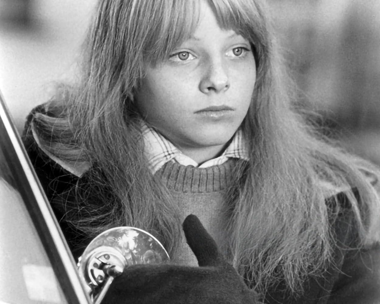 Eine junge Person mit langen Haaren blickt aus der Kamera. Sie trägt einen Pullover, ein Hemd mit Kragen und eine dunkle Jacke. Eine Hand hält ein glänzendes Objekt.