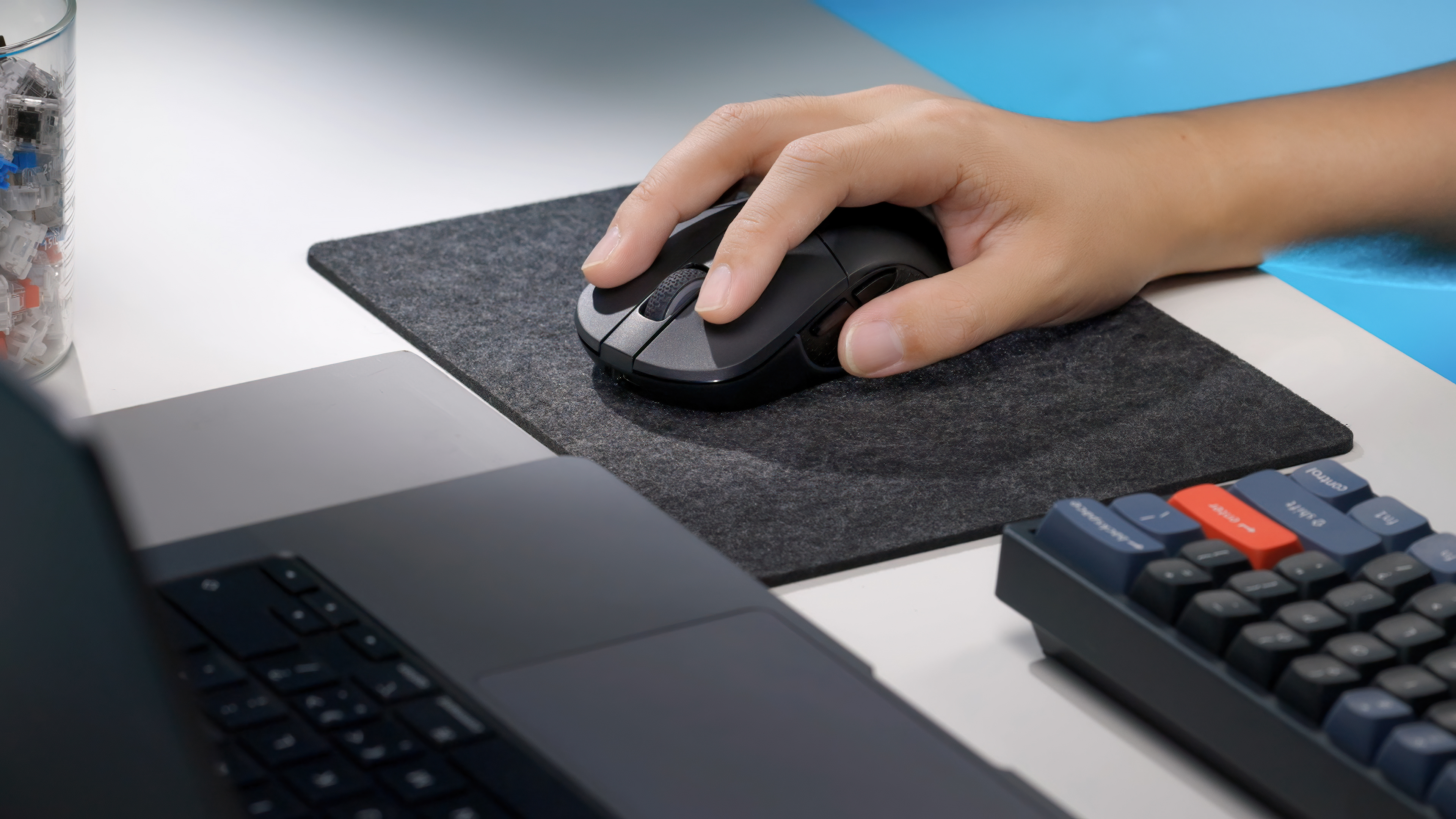 Eine Hand benutzt eine schwarze Maus auf einem grauen Pad. Ein Laptop und eine Tastatur sind ebenfalls vorhanden. Die Szene ist auf einem weißen Schreibtisch.