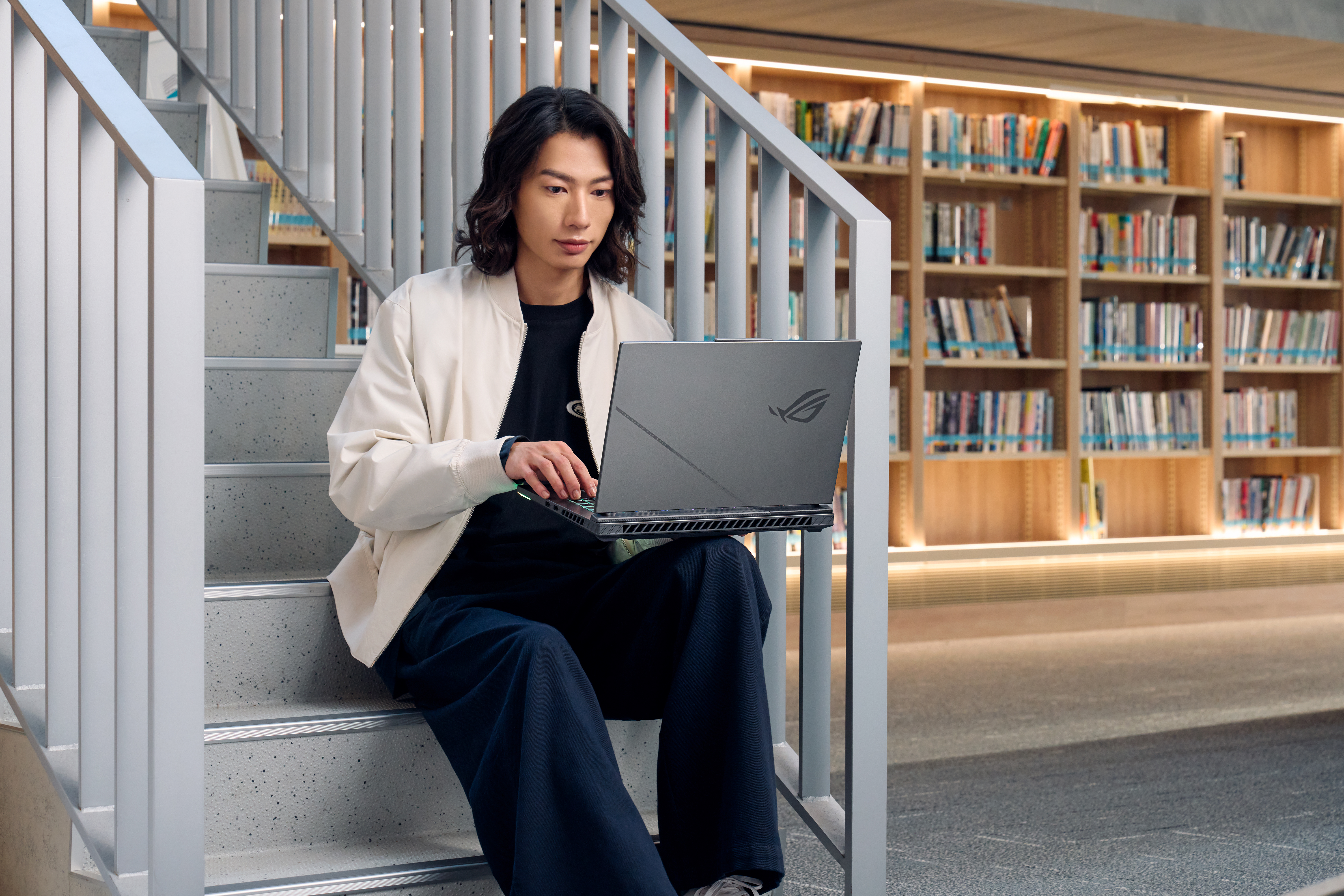 Man zit op trap, laptop gebruikend. Draagt jas en donkere broek. Boekenrekken op de achtergrond.